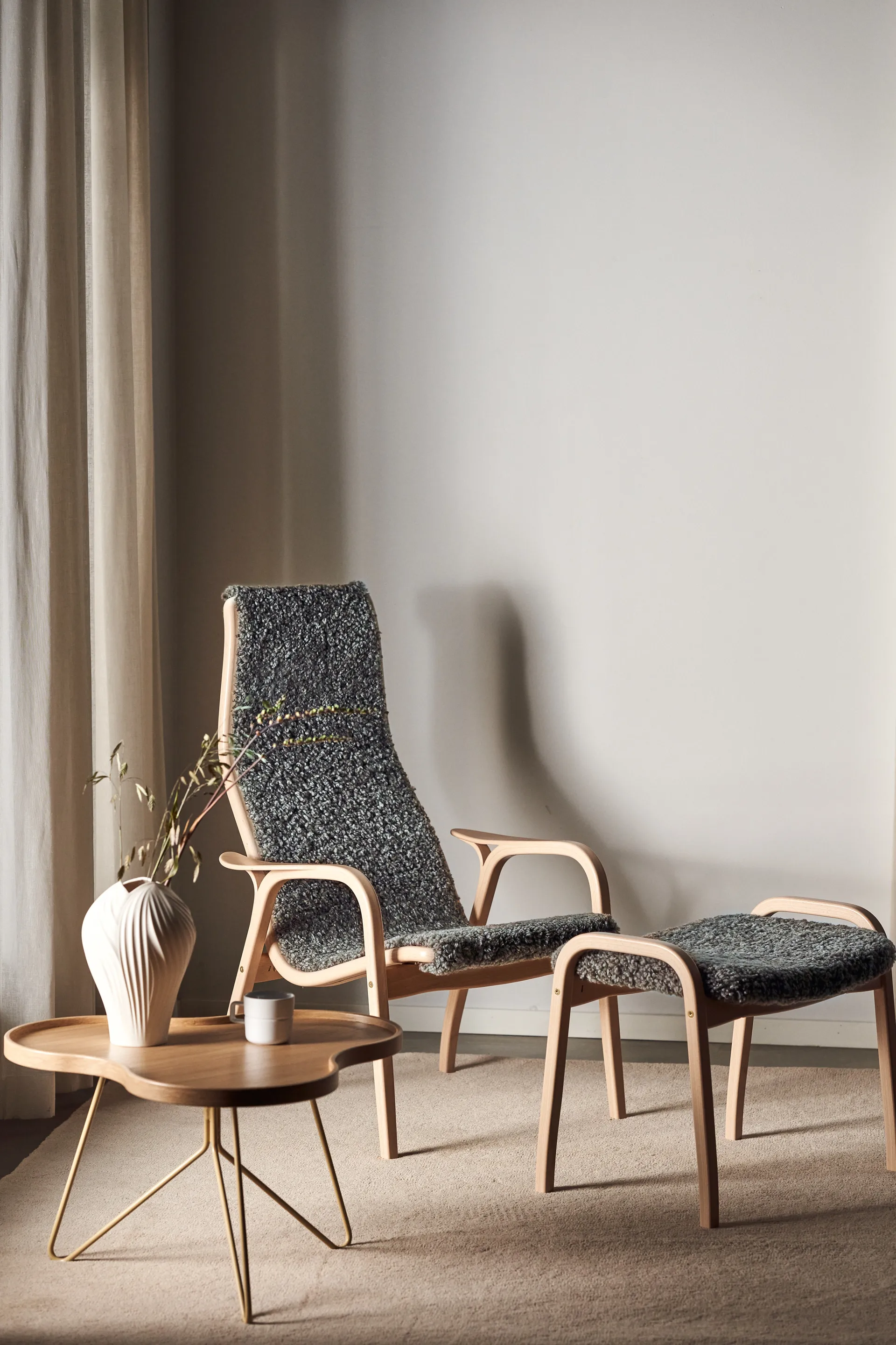 Light wood lounge chair and matching stool with dark gray textured upholstery, next to a small wood coffee table with gold legs, vase, and mug.