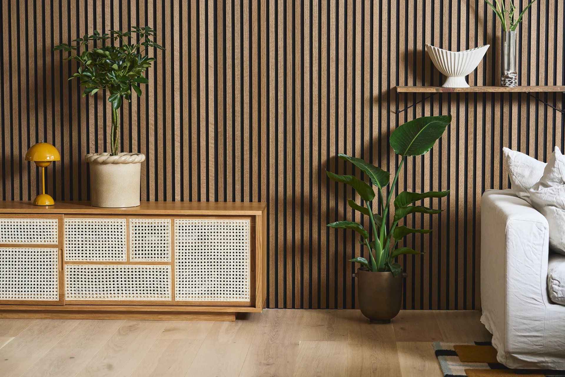 A modern living room with a slatted wooden wall, a rattan cabinet, a yellow lamp, various potted plants, and a white sofa.