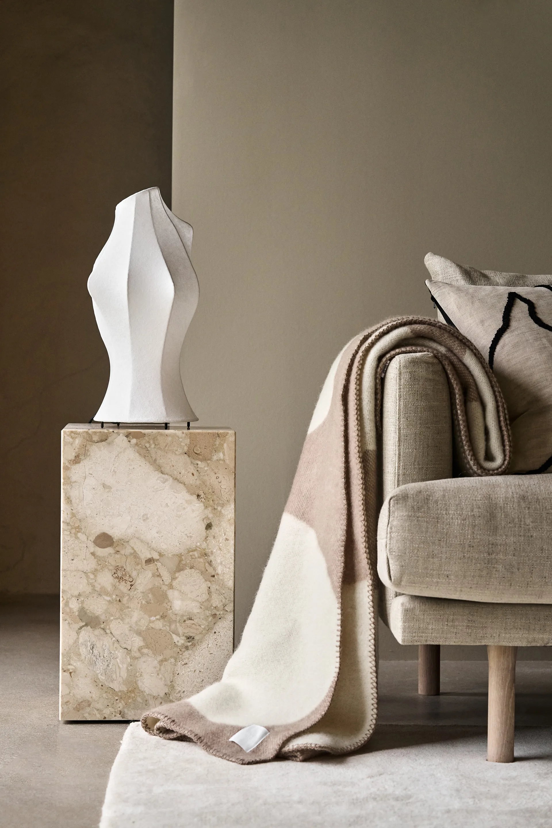 White sculptural lamp on a marble stand, beside a light beige sofa with a folded cream and brown throw blanket and a patterned cushion.