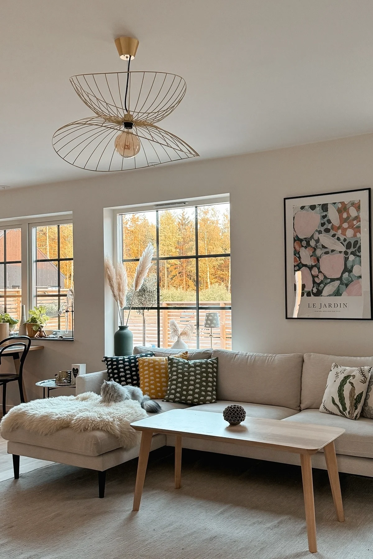 A cozy living room featuring a light sofa, wooden coffee table, unique gold wire pendant light, and windows with autumn tree views. A gray cat rests on a fluffy ottoman.