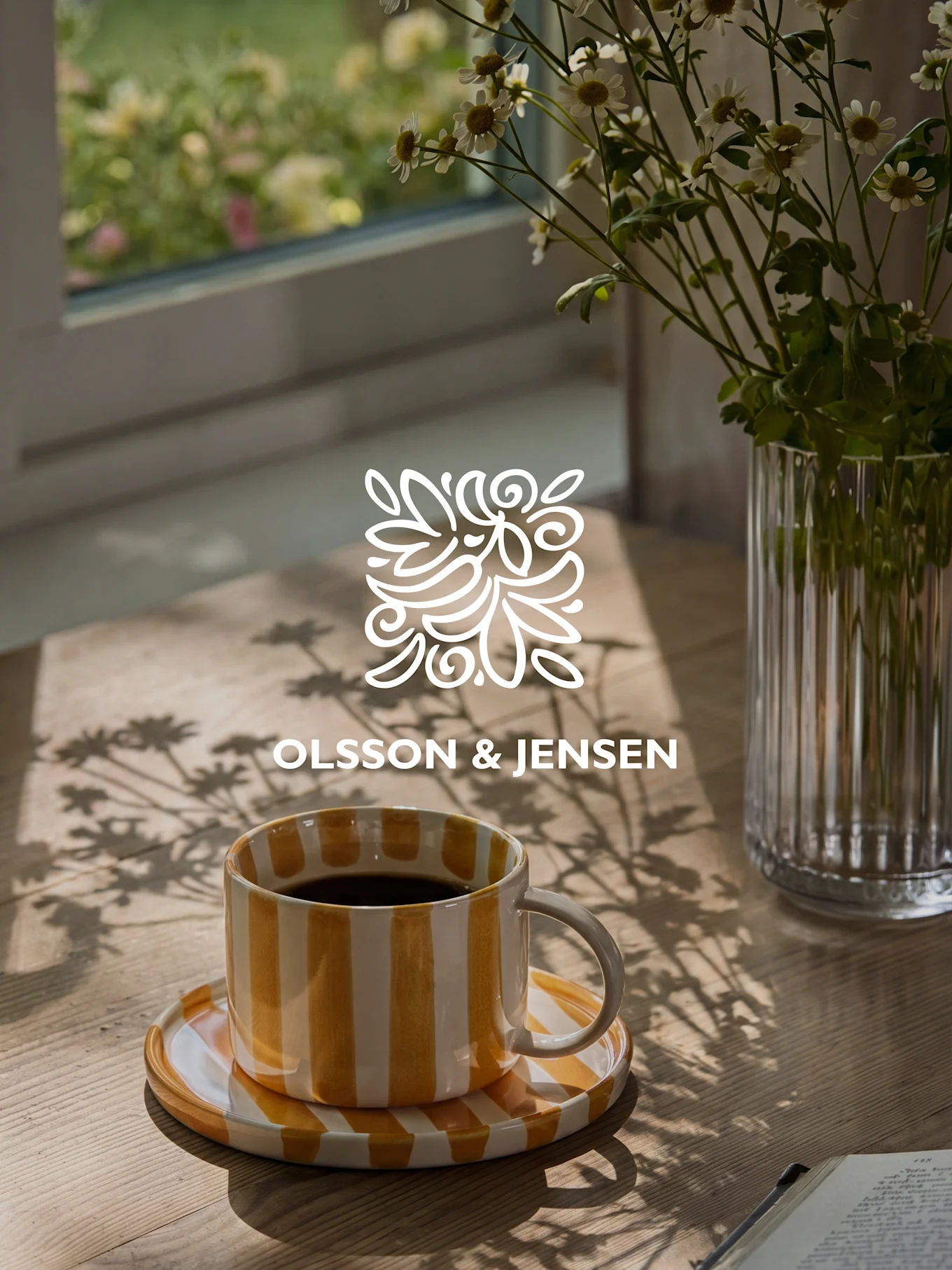 Yellow and white striped coffee mug on a wooden table with a vase of white flowers and sunlit shadows, Olsson & Jensen logo.