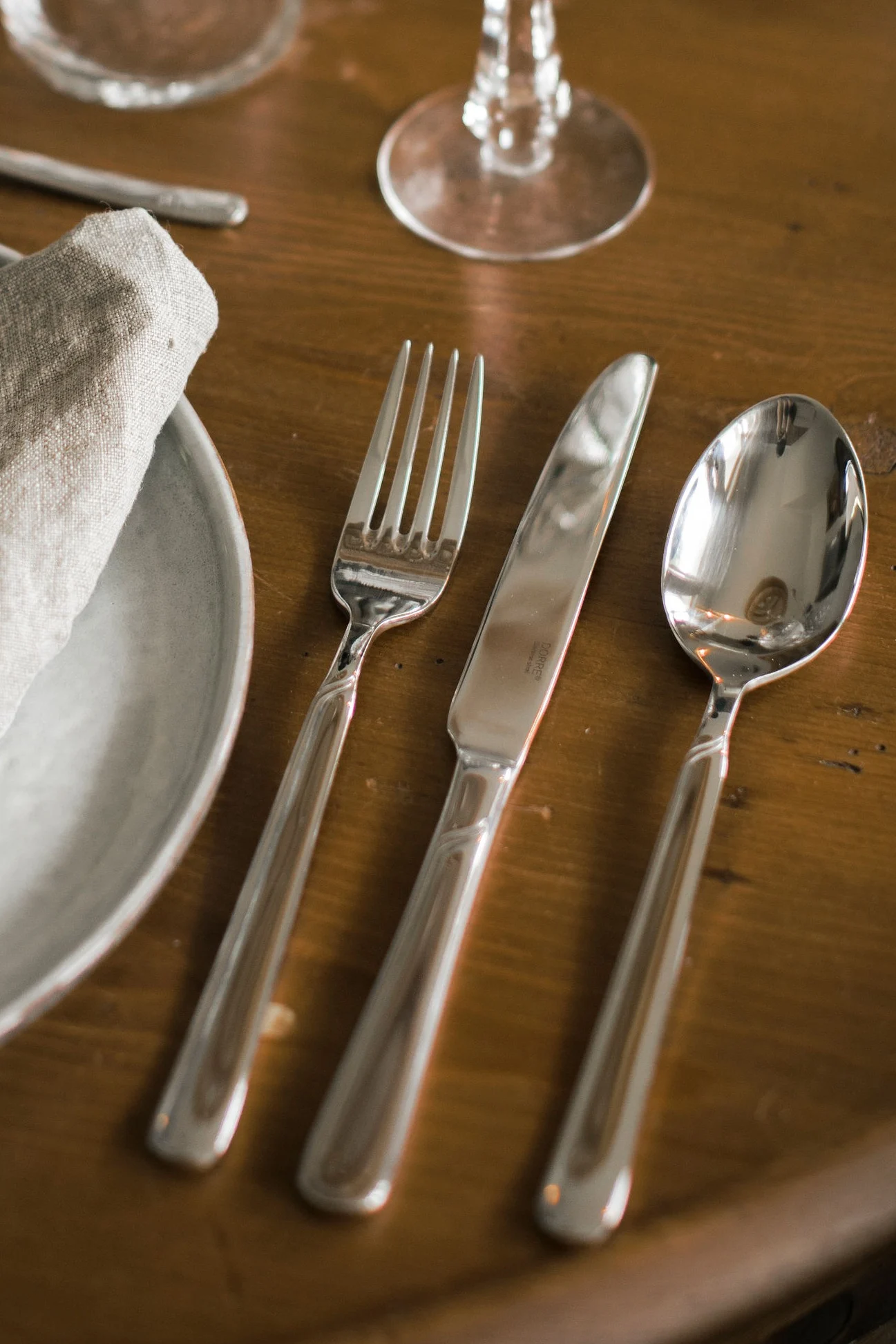 A place setting with a silver fork, knife, and spoon next to a plate with a folded linen napkin on a wooden table.