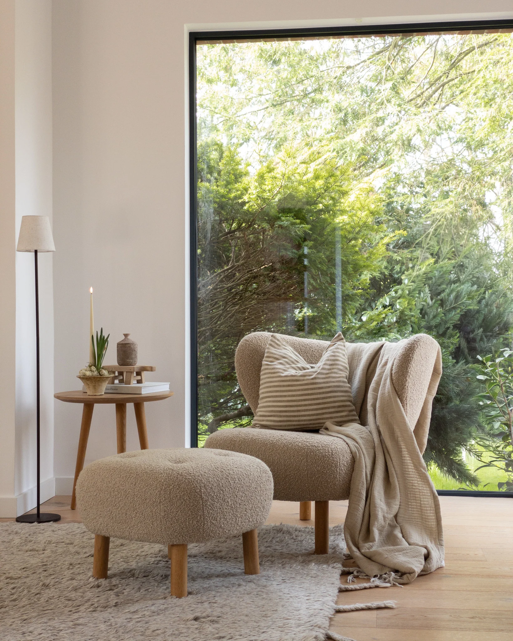 Cozy nook: light brown armchair, ottoman, side table with candle, large window, green trees.