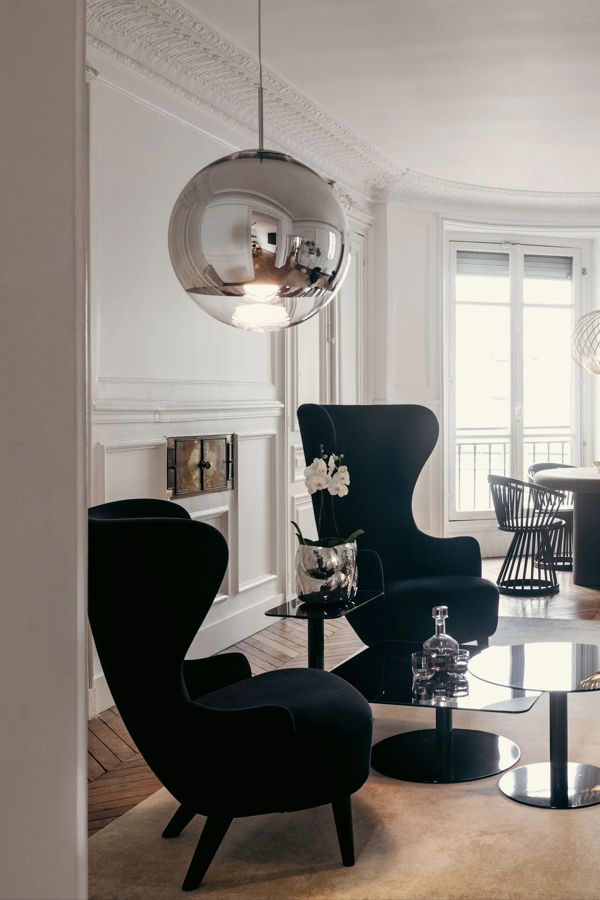 Elegant living room with two black armchairs, glass tables, and a large spherical silver pendant lamp.