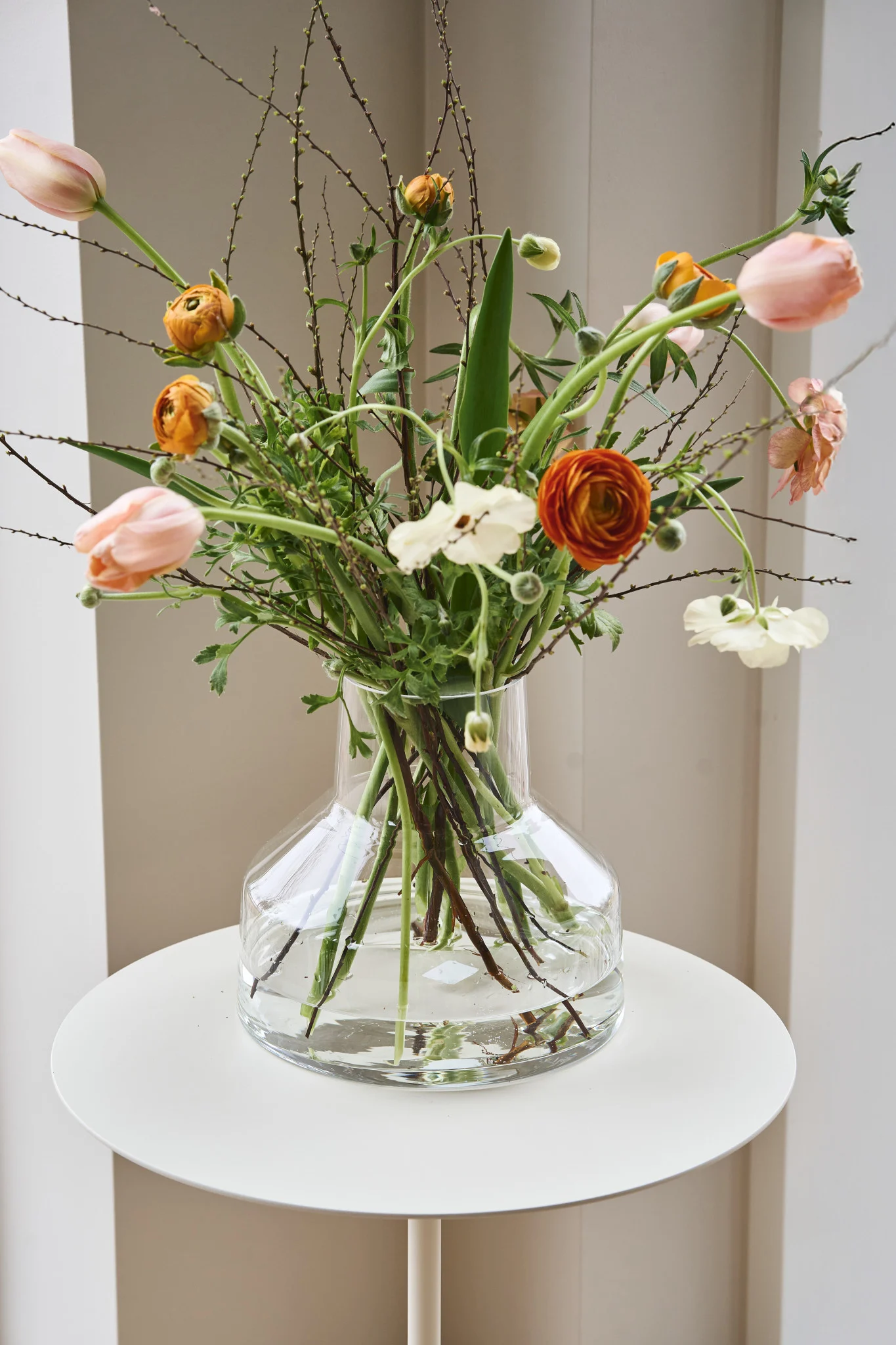 A vibrant floral arrangement in a clear glass vase with peach tulips, orange and white ranunculus, and budding branches on a white table.