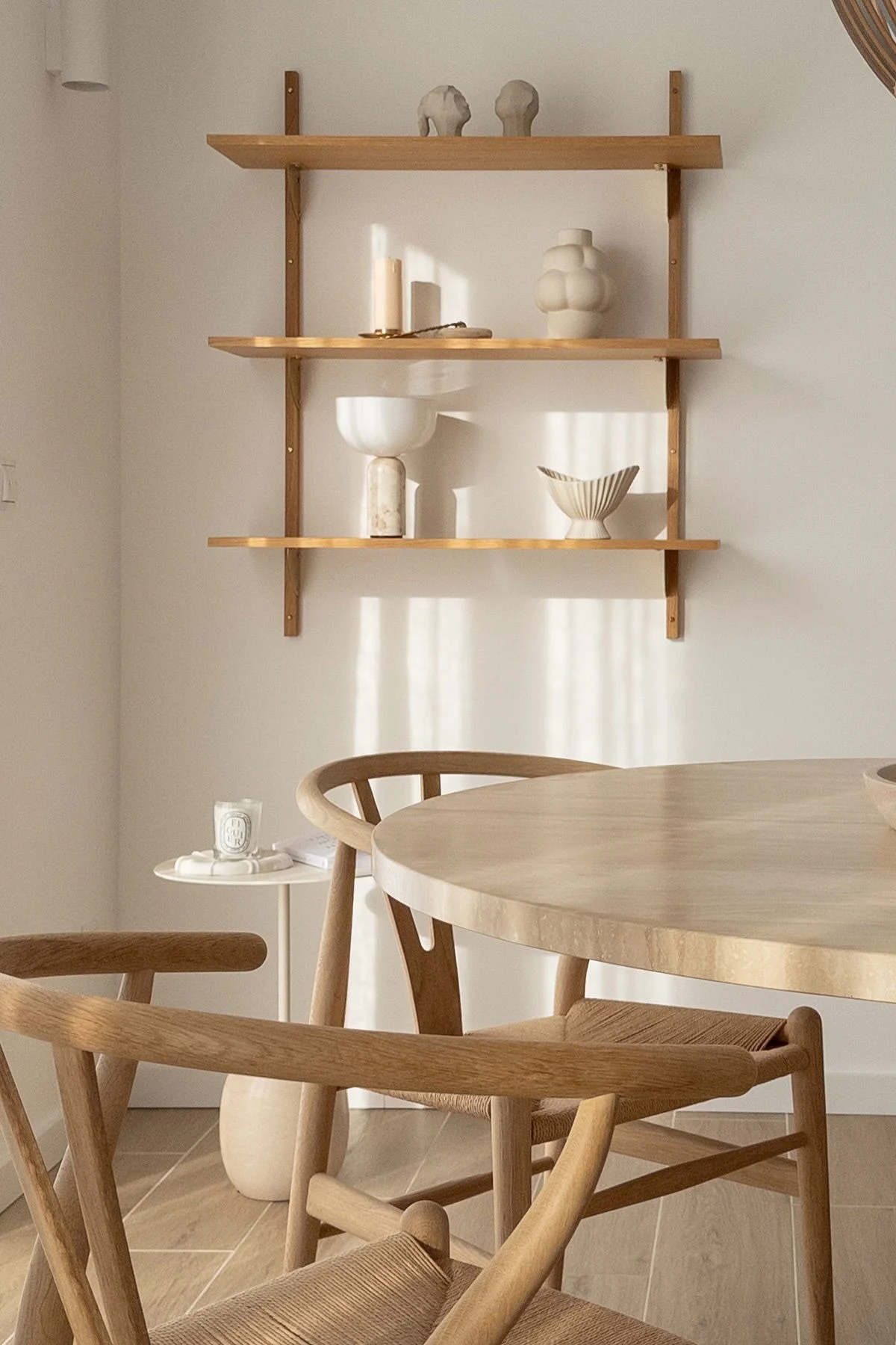 Minimalist dining area with a round light wood table and chairs with woven seats. Wooden shelves display decorative ceramics and candles.