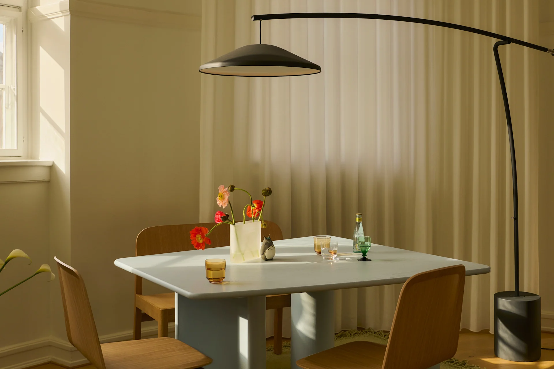 A modern dining room with a light blue table, wooden chairs, a black arched floor lamp, and a vase of colorful flowers in sunlight.