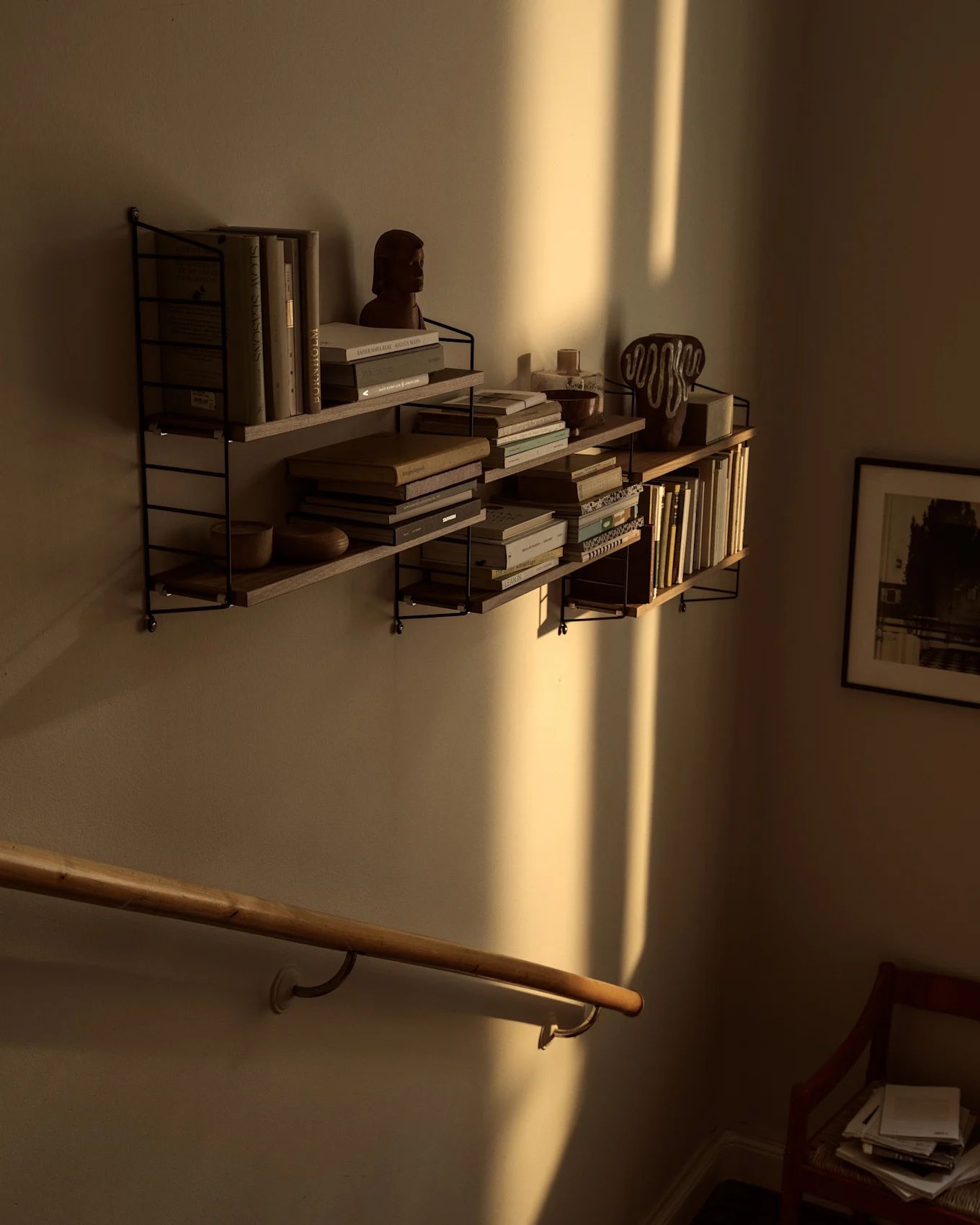 Wall-mounted shelves filled with books and decor, bathed in warm sunlight casting strong vertical shadows above a wooden handrail.