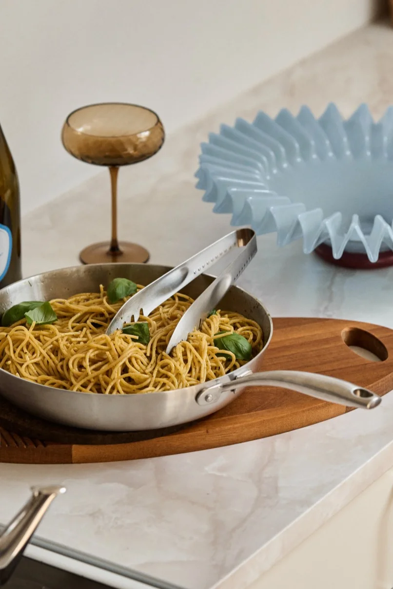 Spaghetti with basil in a metal pan with tongs on a wooden board, next to a wine bottle, glass, and a decorative blue bowl.