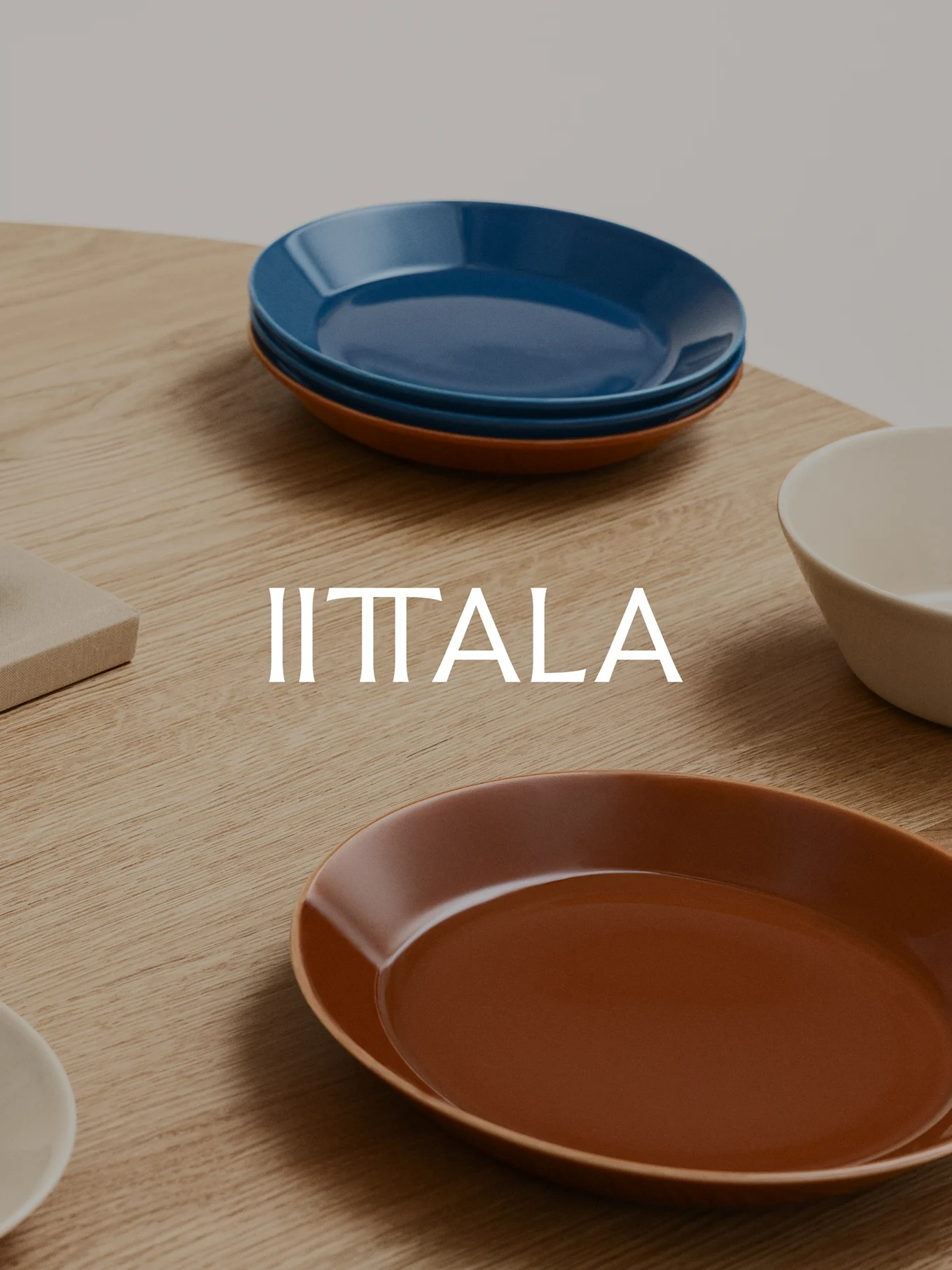 Iittala logo with a stack of blue plates, a brown plate, and a beige bowl on a wooden table.