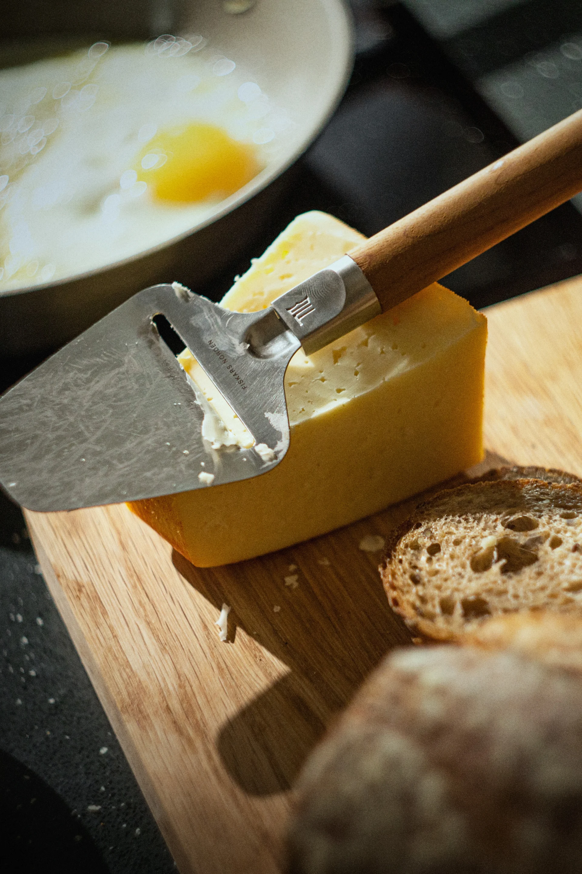 A cheese slicer rests on a block of yellow cheese on a wooden board, with bread slices and a fried egg in a pan in the background.