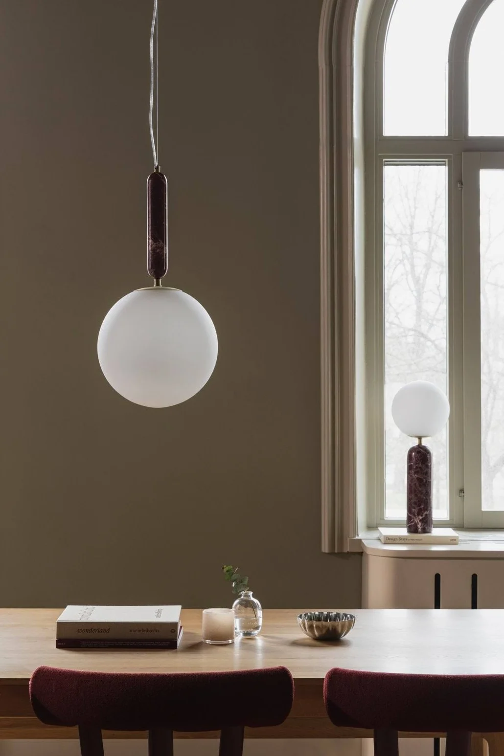 A dining room with a wooden table and maroon chairs, featuring a spherical pendant light and a matching table lamp with dark red marble accents.