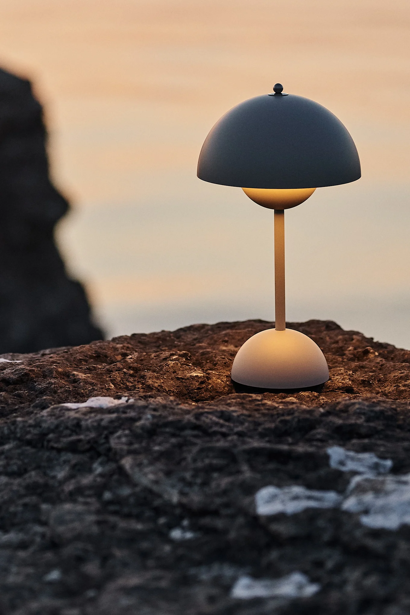 A modern table lamp with a dark domed shade and glowing light-colored stem sits on a textured rock at sunset.