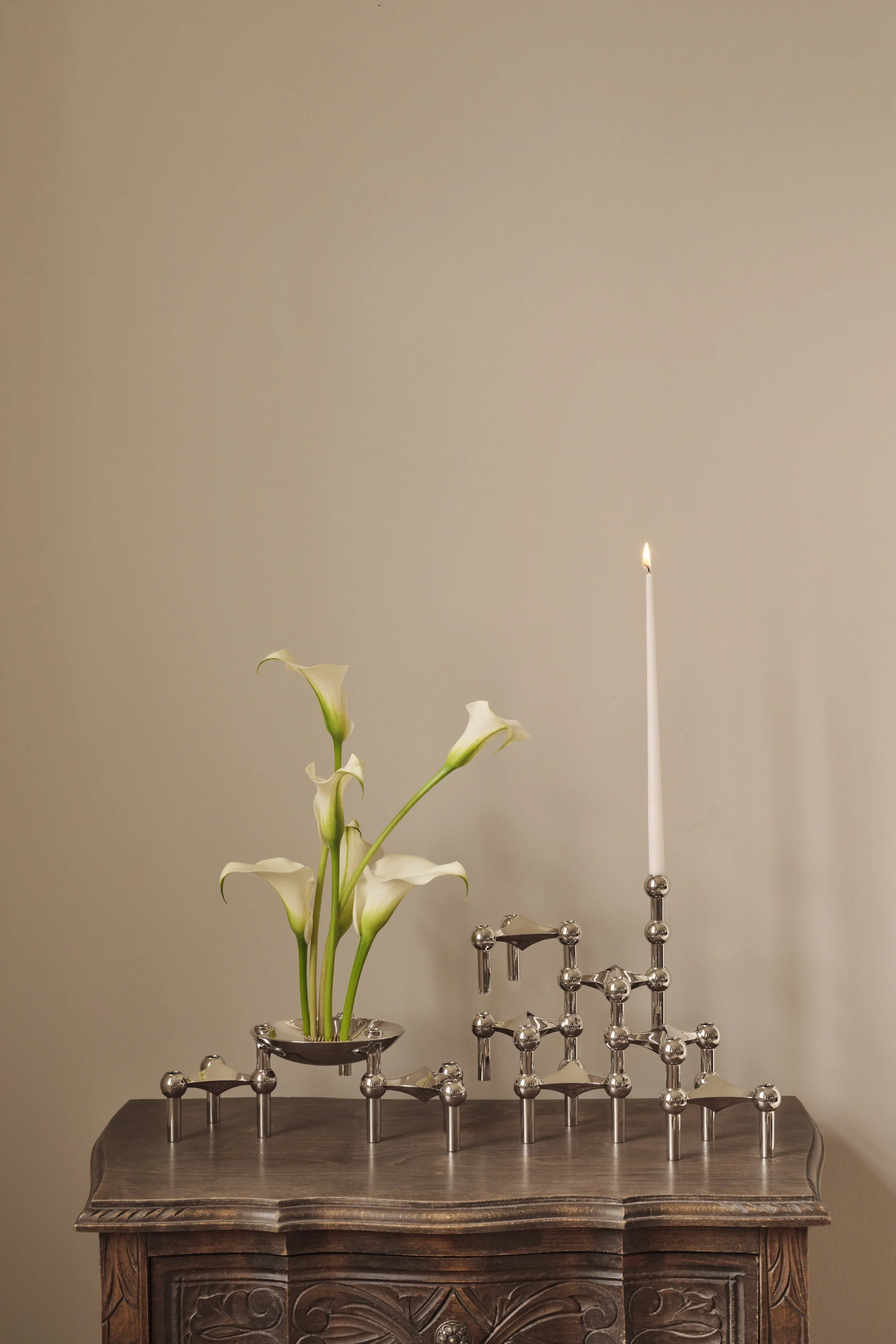 White calla lilies in a silver bowl next to a modular silver candle holder with one lit white candle on a carved wooden table.