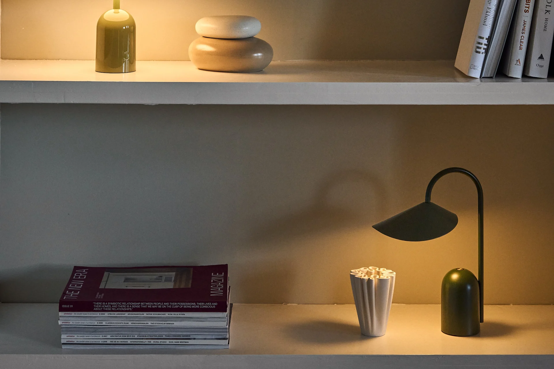 Three shelves display various desk lamps in green and purple, alongside books, magazines, and decorative objects.