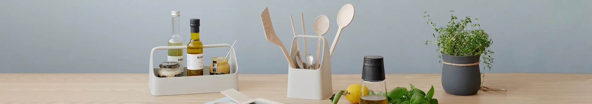Kitchen essentials on wood table: caddy with oils and jars, utensil holder with wooden tools, olive oil, lemon, basil, potted herb.