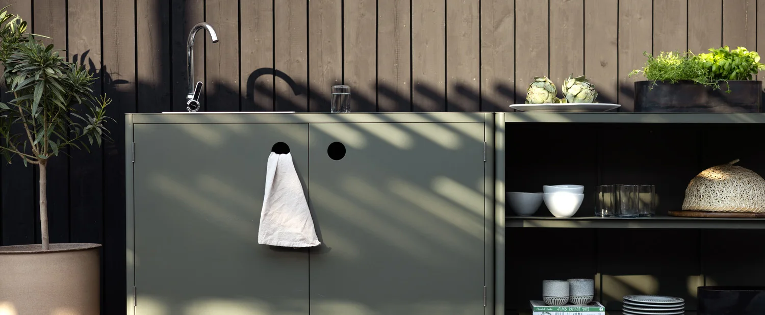 Outdoor kitchen with sage green cabinets, sink, herbs, and a potted plant against a dark wooden wall.
