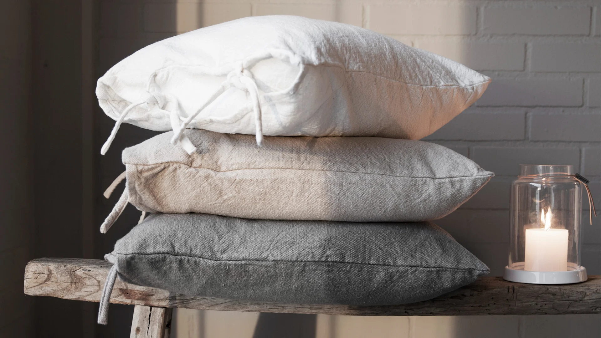 Stack of three linen pillows (white, beige, grey) on a rustic wooden bench next to a lit candle in a glass lantern.