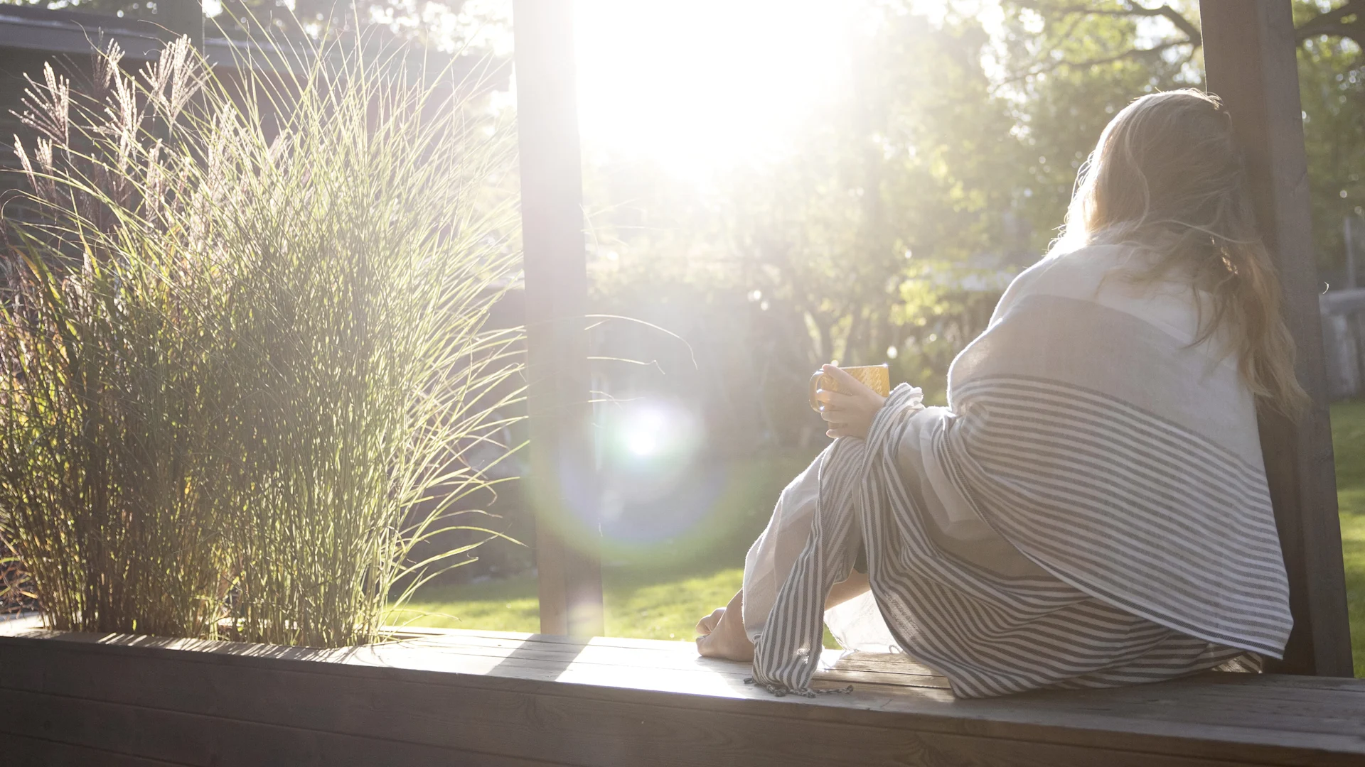 Person wrapped in a striped blanket, holding a mug and looking at a sunny outdoor scene with green grass and trees.