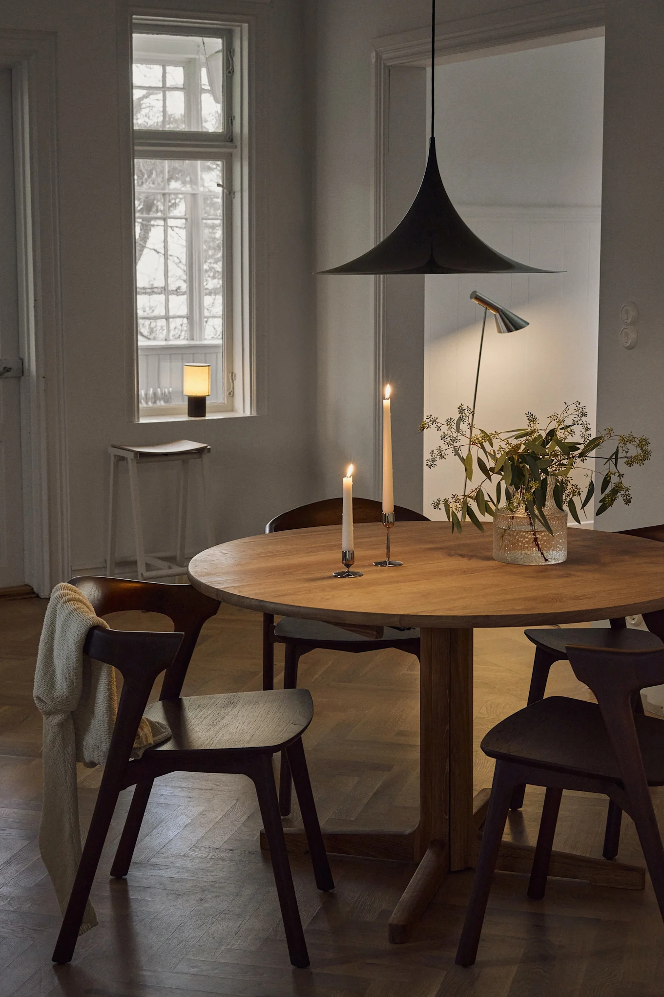 Dining room with a round wooden table, chairs, lit candles, a vase with greenery, and various lamps, featuring a large pendant light.