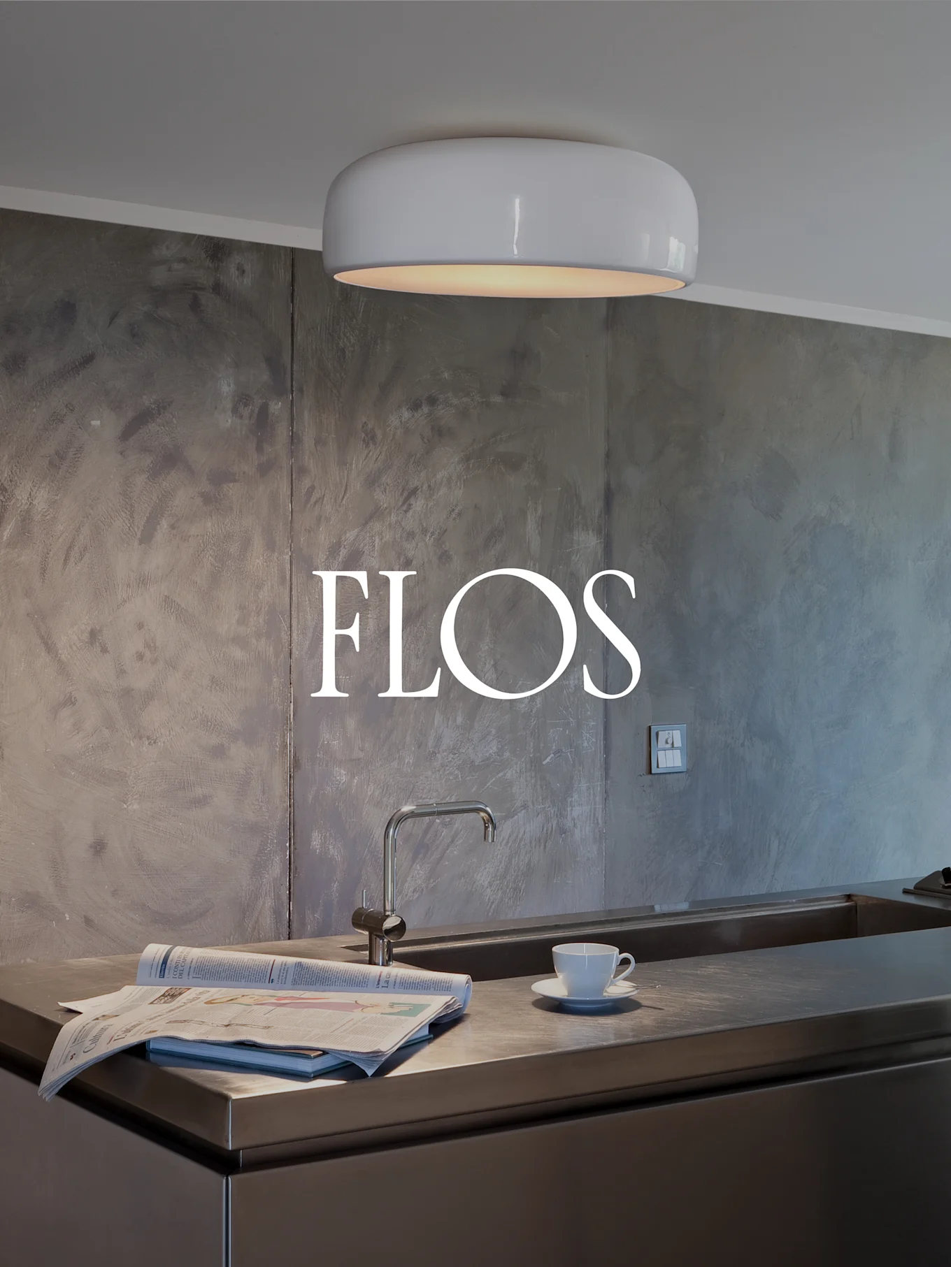 Modern kitchen with a white, rounded ceiling light, a stainless steel counter with sink, newspaper, and teacup. FLOS logo on textured wall.