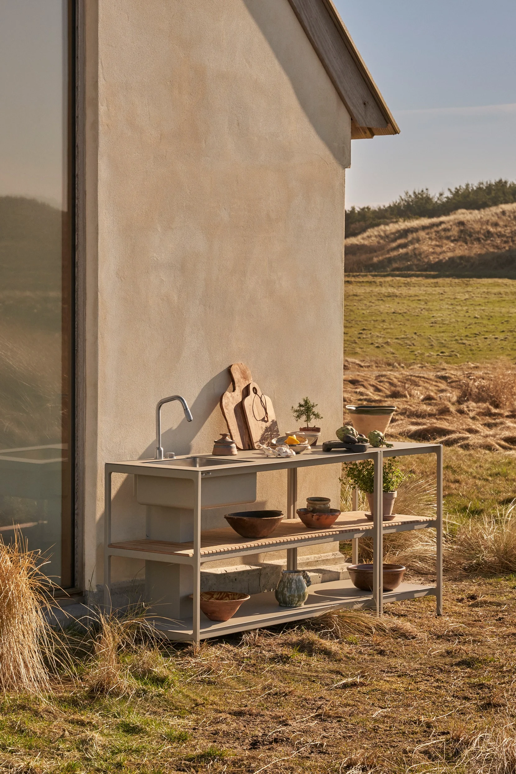 Outdoor kitchen unit with sink and shelves, styled with cutting boards, plants, and bowls, against a building in a natural landscape.