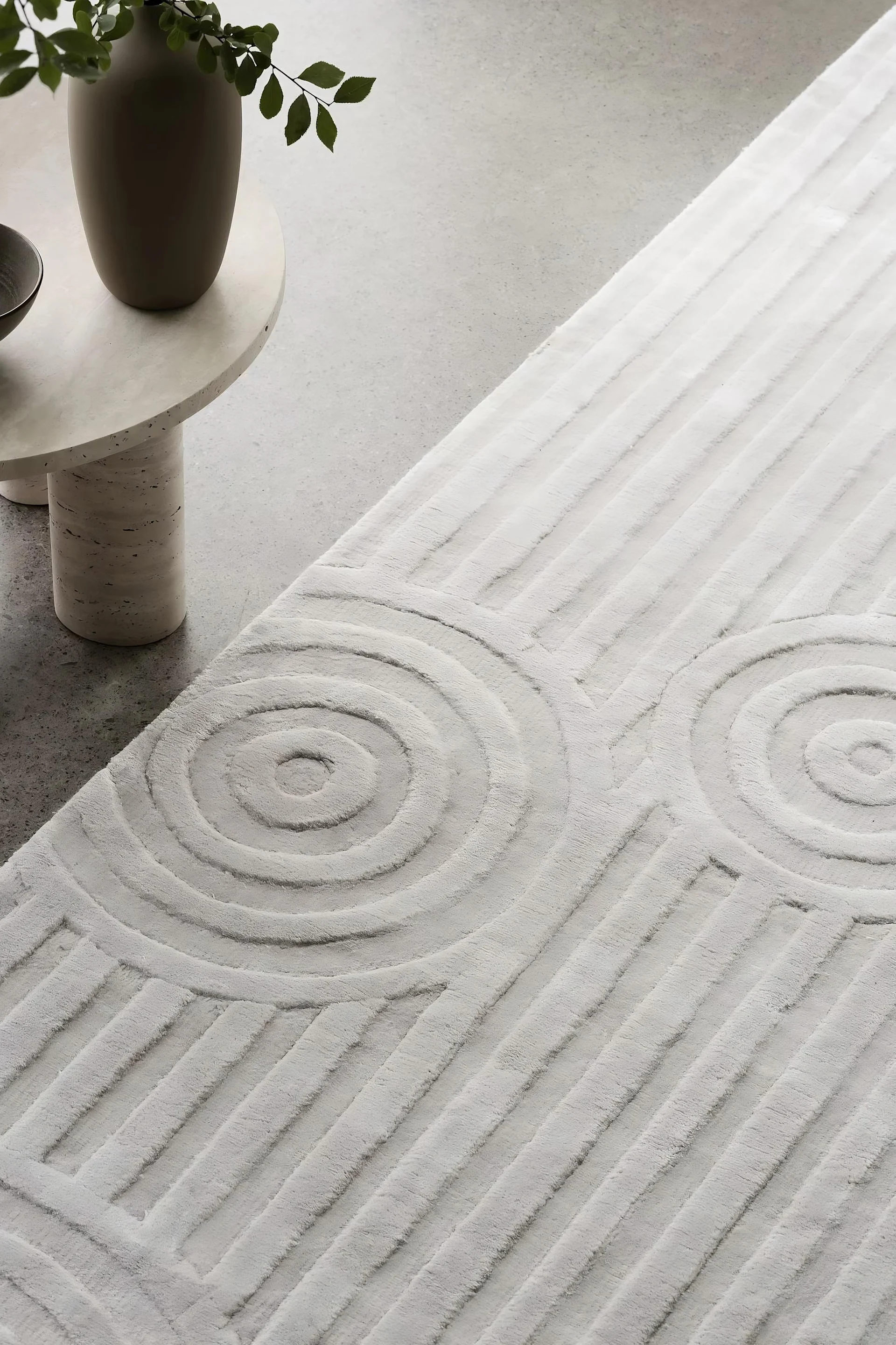 Textured light grey rug with concentric circle and line patterns on concrete, next to a small table and vase.