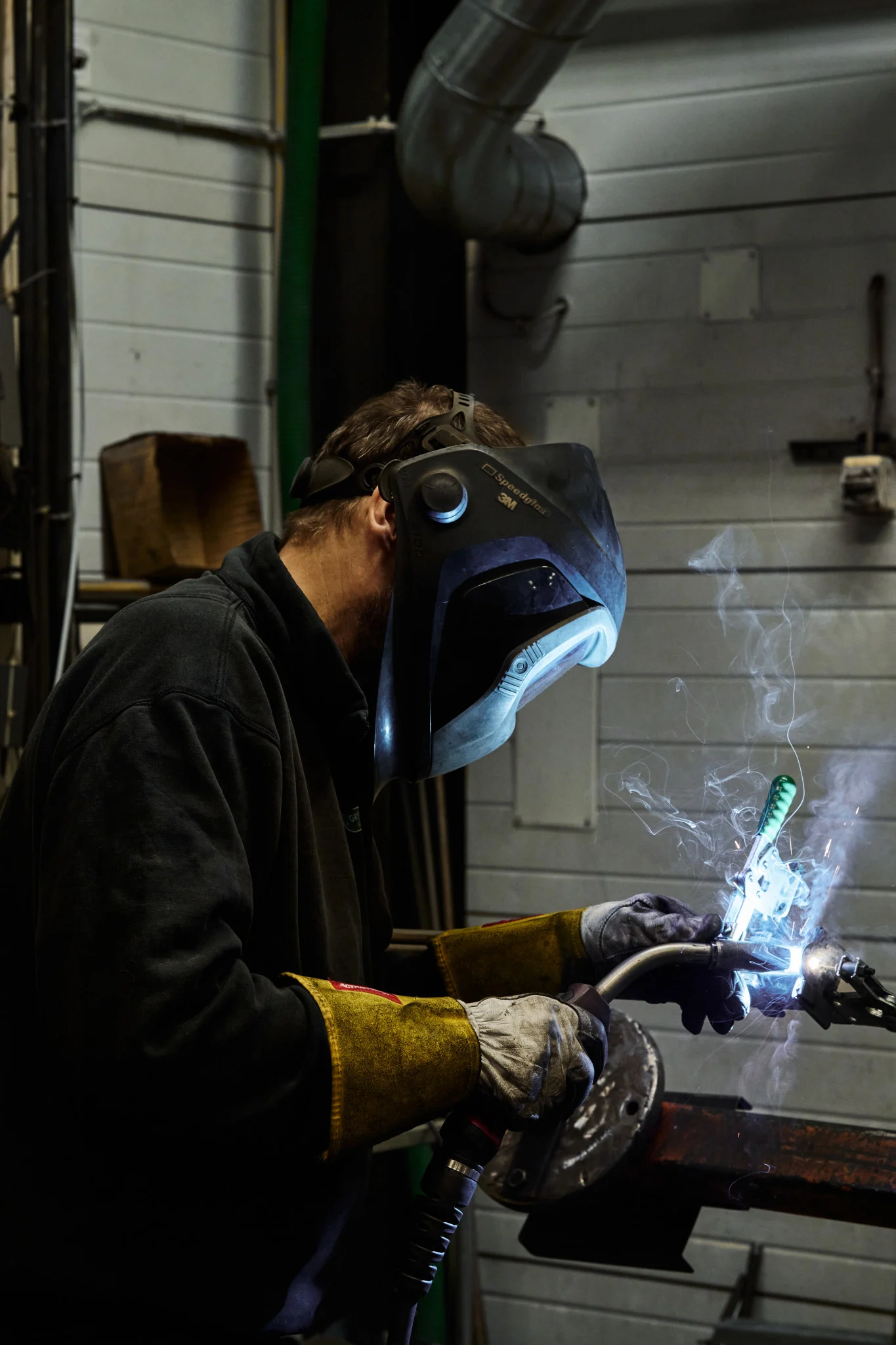 A person in a welding helmet and gloves performs welding, with bright sparks and smoke visible.