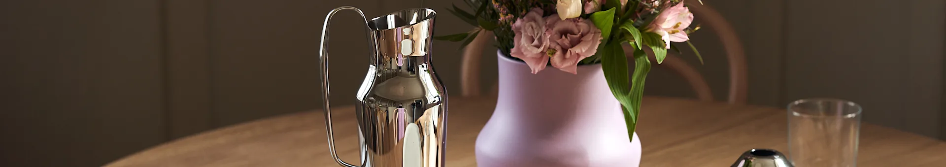 Polished silver carafe next to a light purple vase with pink flowers on a wooden table.