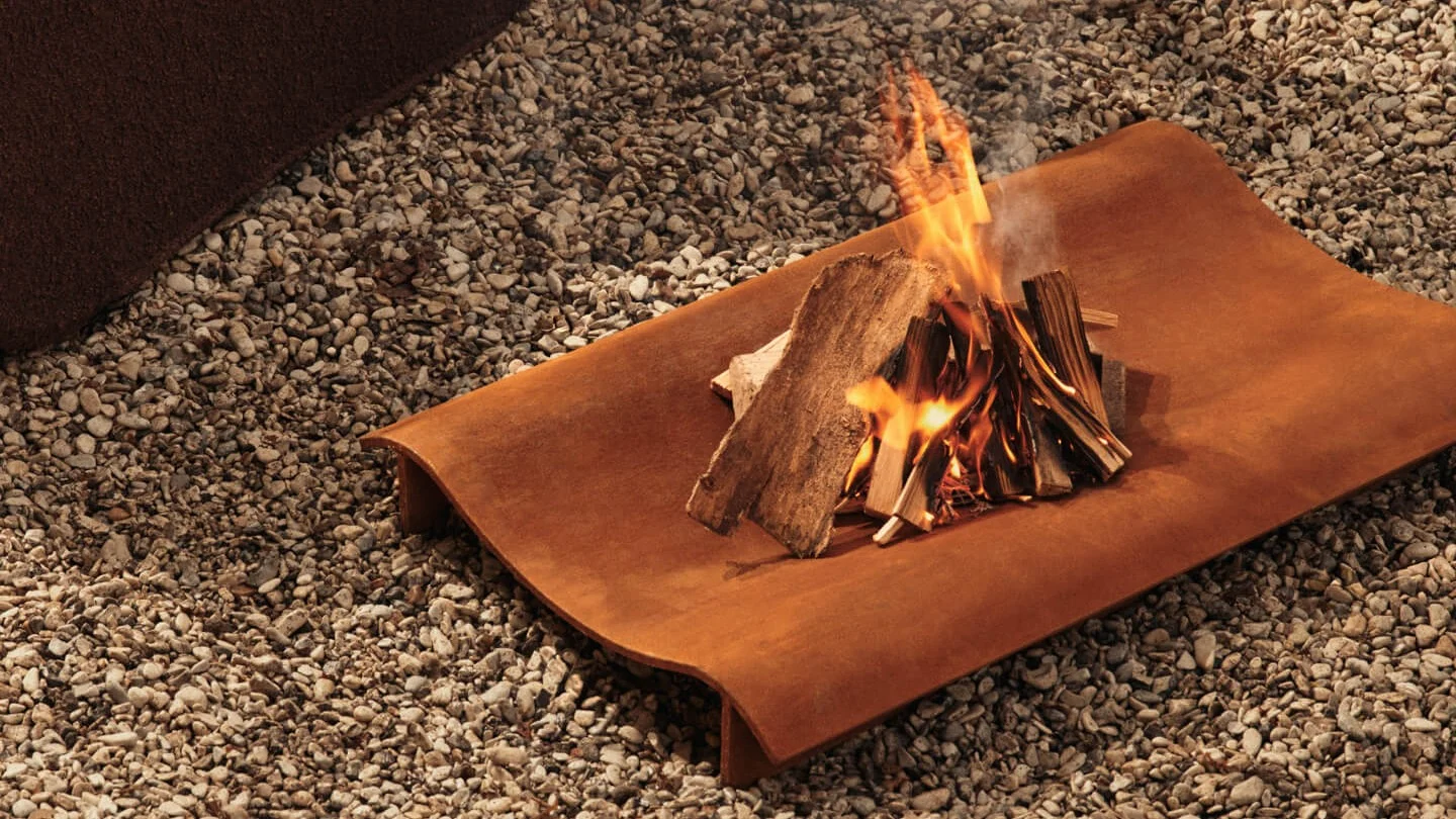 Small bonfire with burning wood on a rectangular, rusted metal tray over gravel.