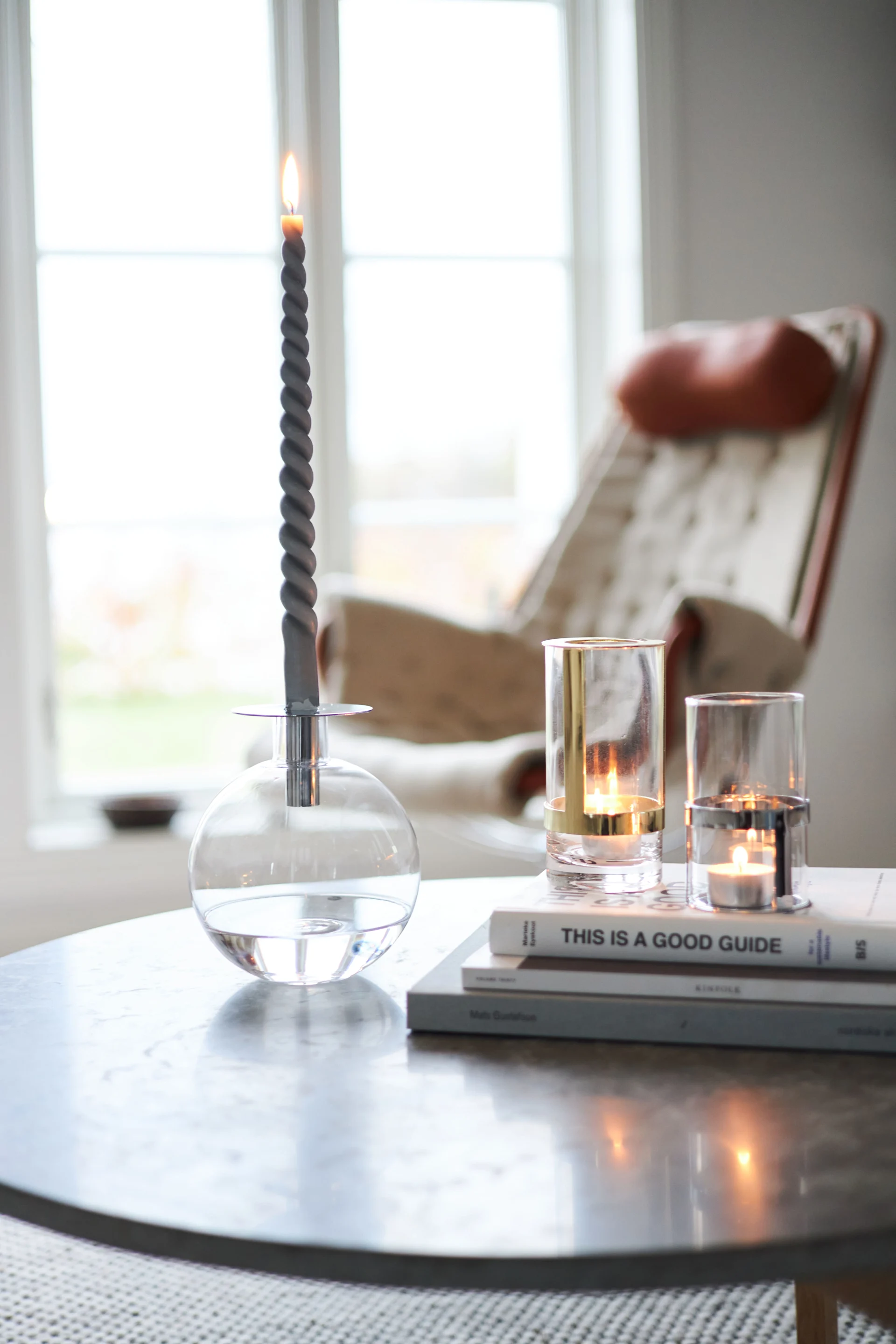 Lit grey twisted candle in a glass holder, two glass tealight holders on books, on a table with a bright window and armchair.
