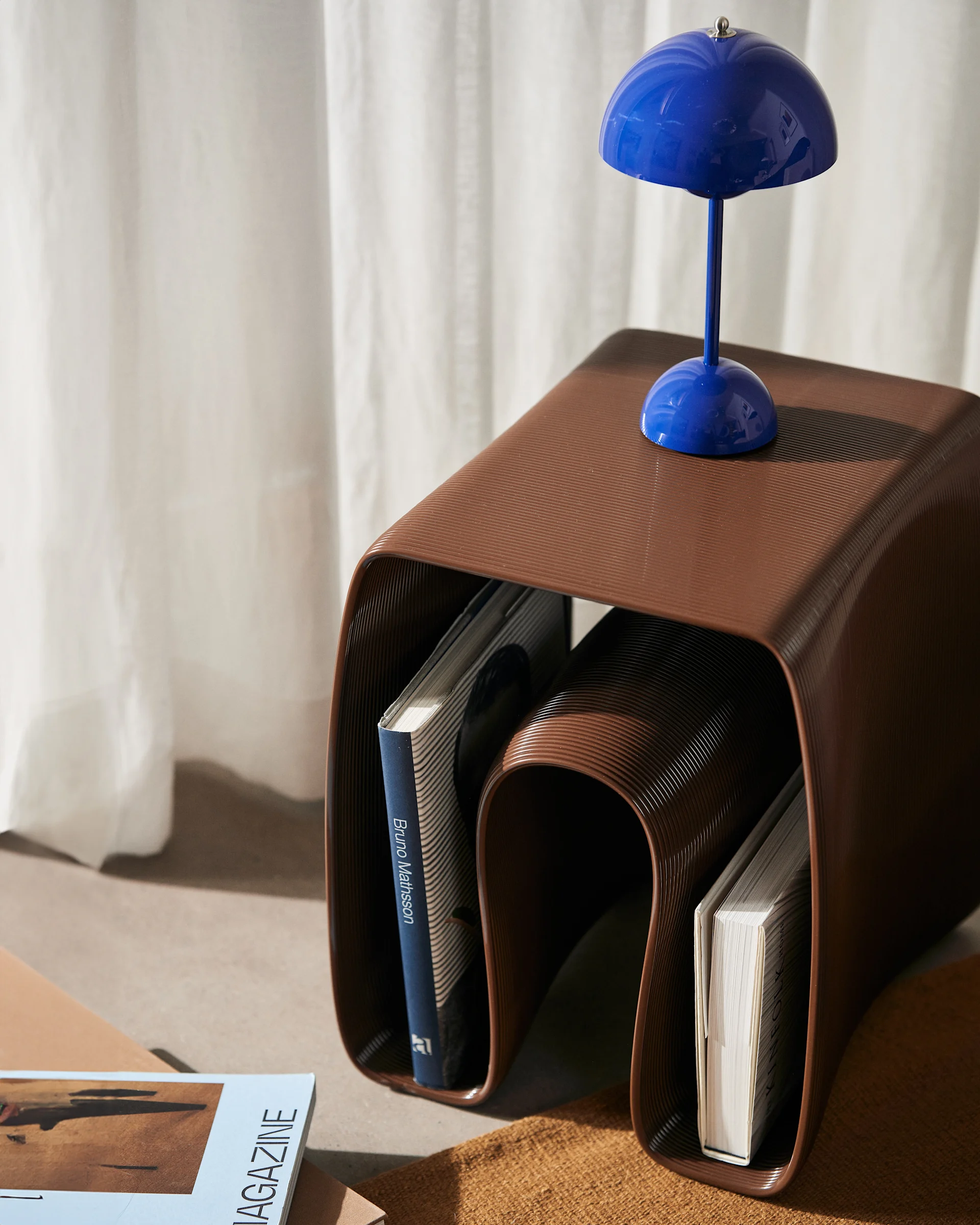 Brown corrugated side table with books, topped by a blue mushroom lamp against a white curtain.
