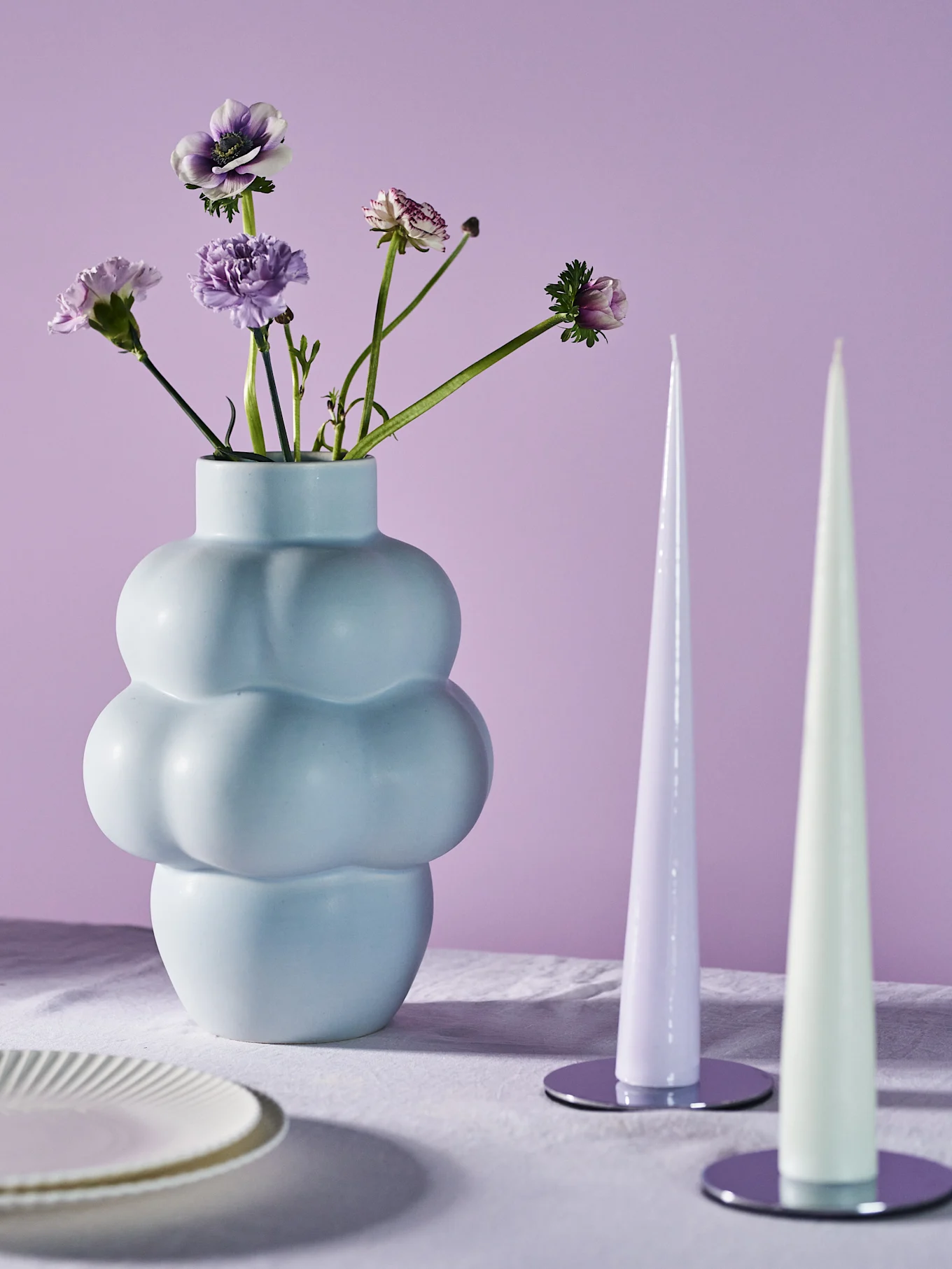 A light blue, bulbous vase with purple and white flowers, and two tapered candles on a white tablecloth.
