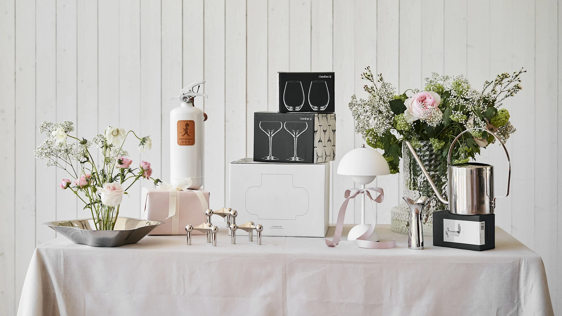 Table with flower bouquets, a white extinguisher, boxed glassware, silver candle holders, a white lamp, and a watering can.