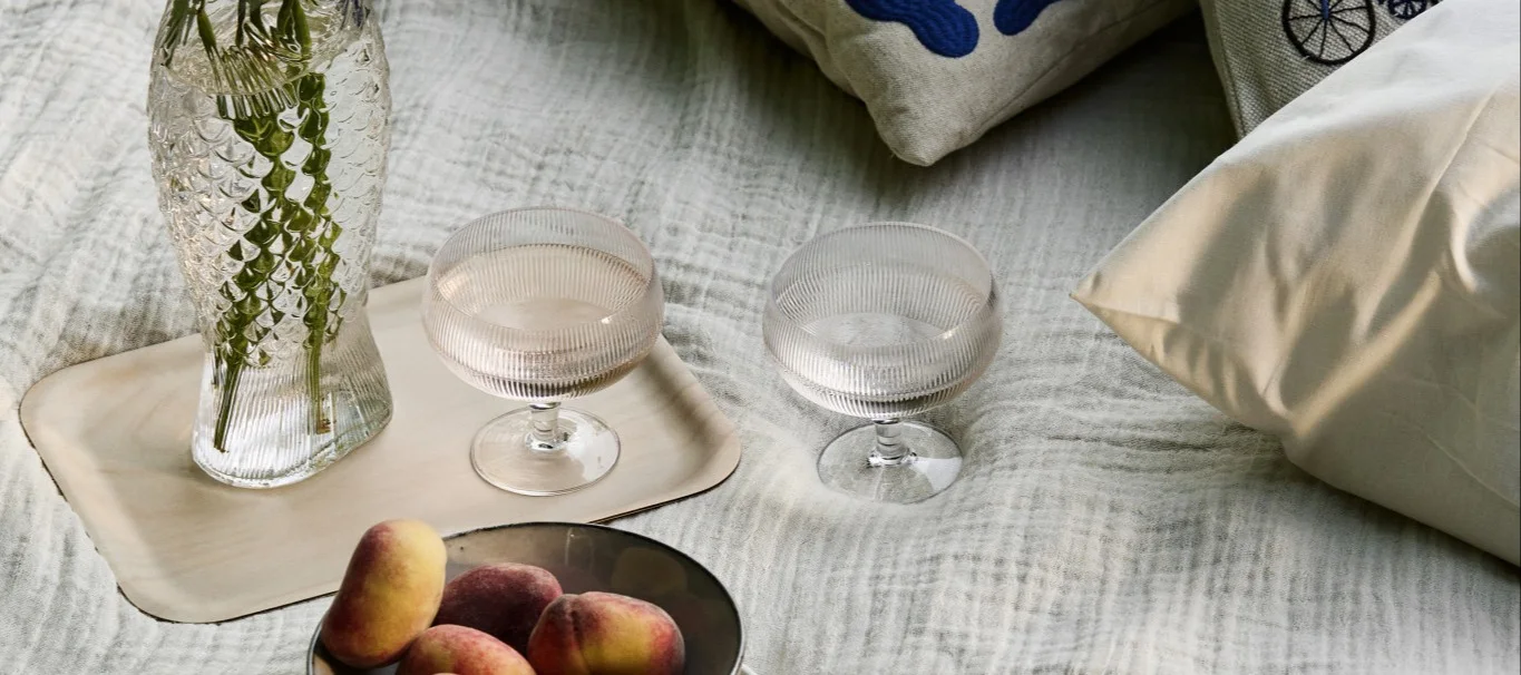 Picnic setup with a fish-shaped vase, two ribbed goblets, and peaches on a light blanket.