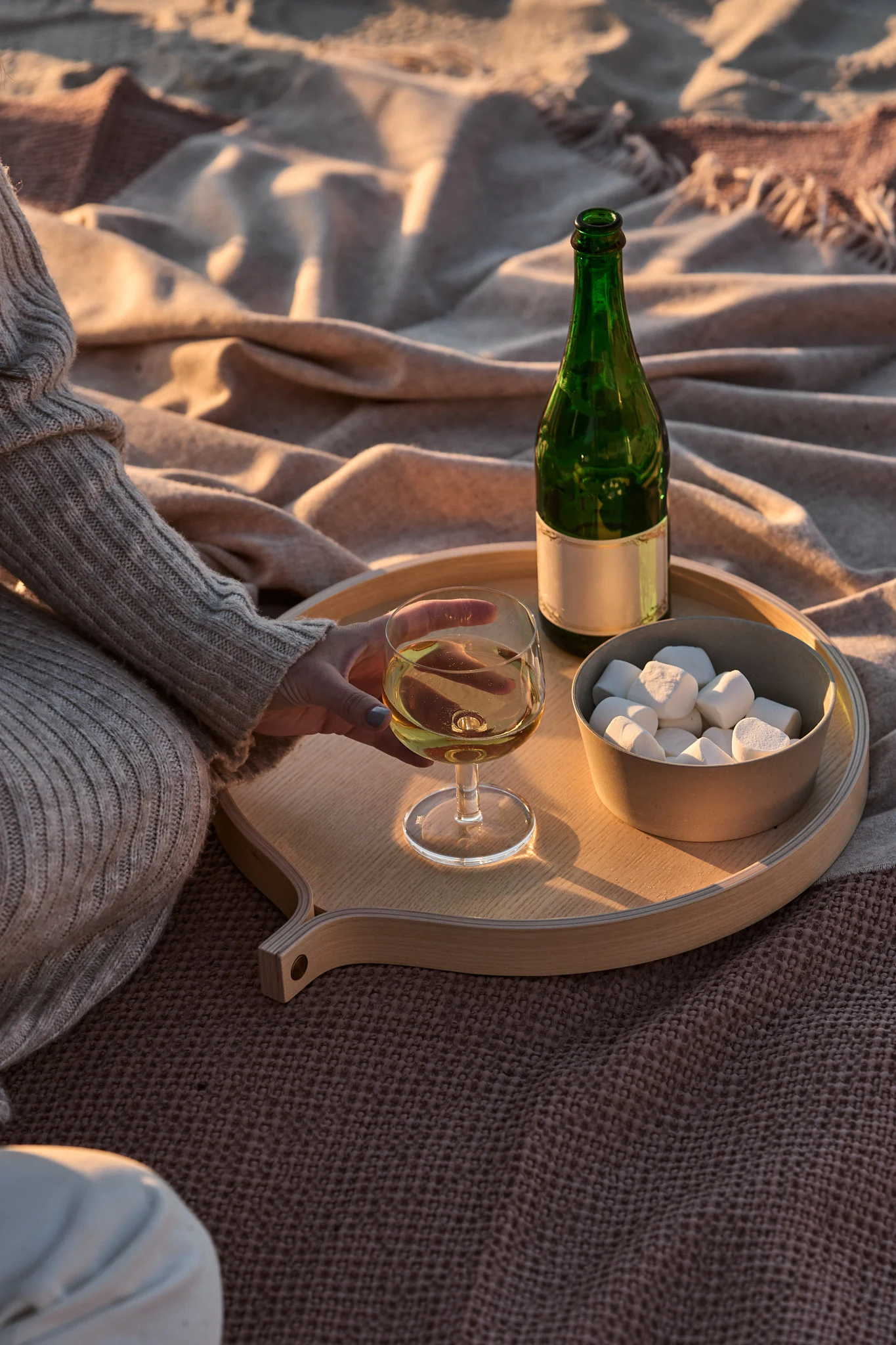 Person holding white wine on a wooden tray with a green bottle and marshmallows on blankets during golden hour.