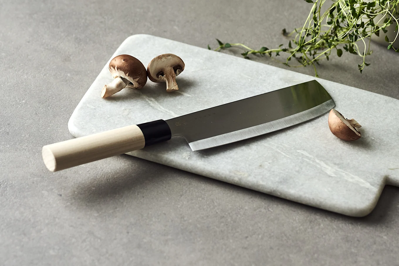 Chef's knife with wooden handle on marble board, next to whole and sliced brown mushrooms and a sprig of thyme.