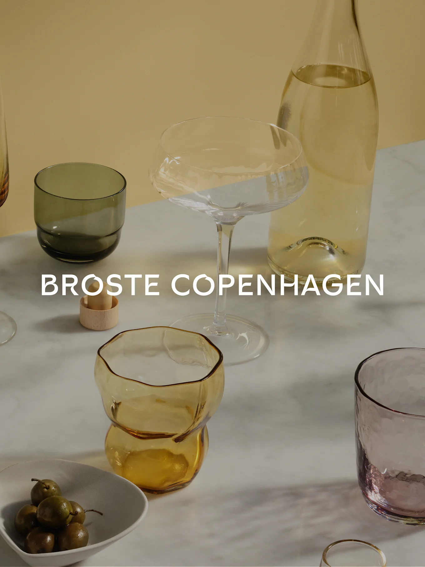 Glassware assortment: clear bottle, coupe, tumblers in olive green, amber, and purple on marble, with olives. Broste Copenhagen.