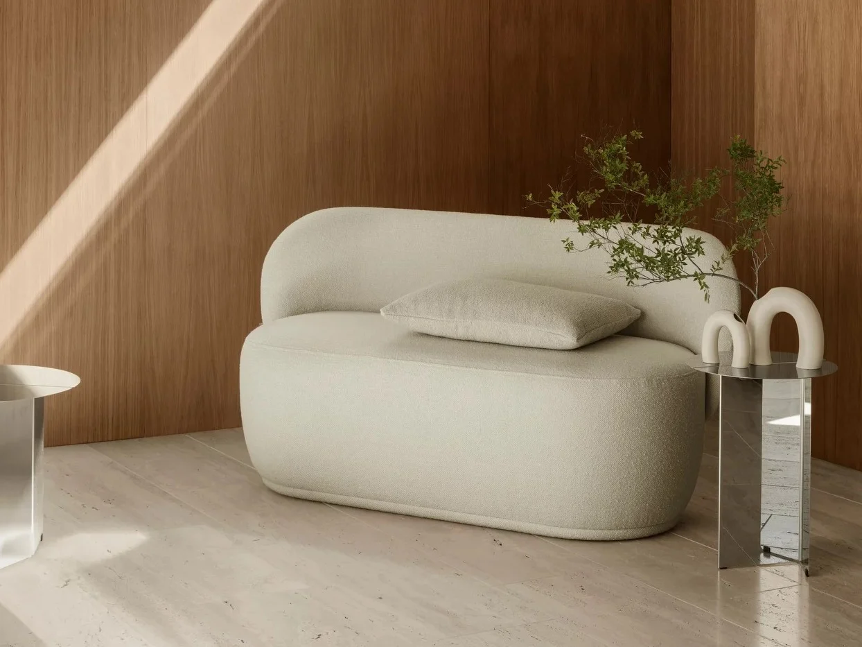 Cream rounded two-seater sofa with pillow on light floor, wood panel wall. Mirrored table with U-shaped decor, leafy plant.