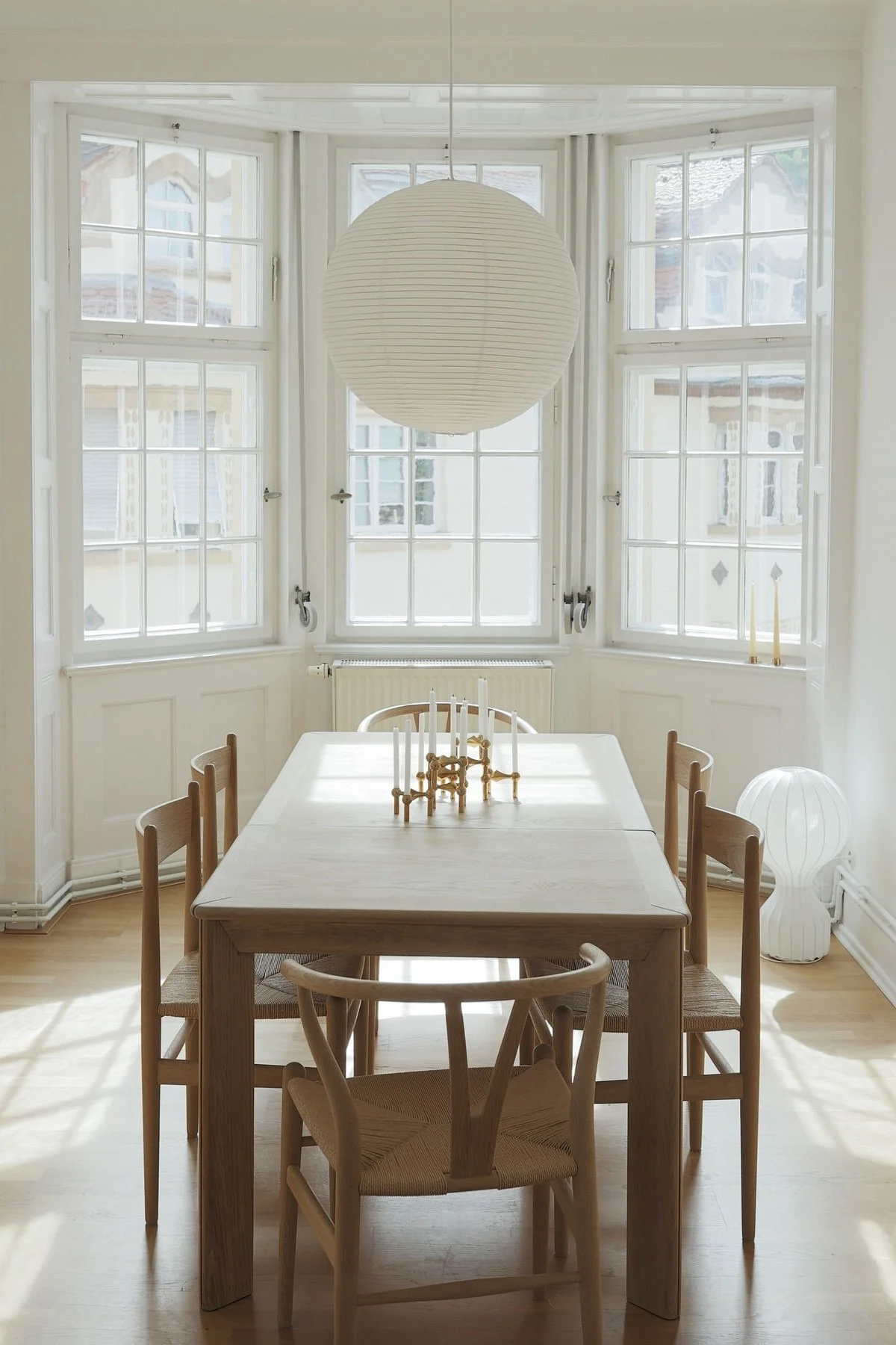 Bright dining room with a light wood table, six chairs, a large paper lantern, and a gold candlestick holder on the table. Sunlight streams through bay windows.