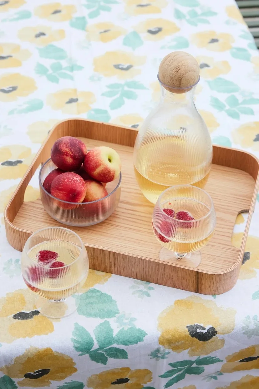 A wooden tray with a glass pitcher, two glasses of raspberry spritzers, and a bowl of nectarines on a floral tablecloth.