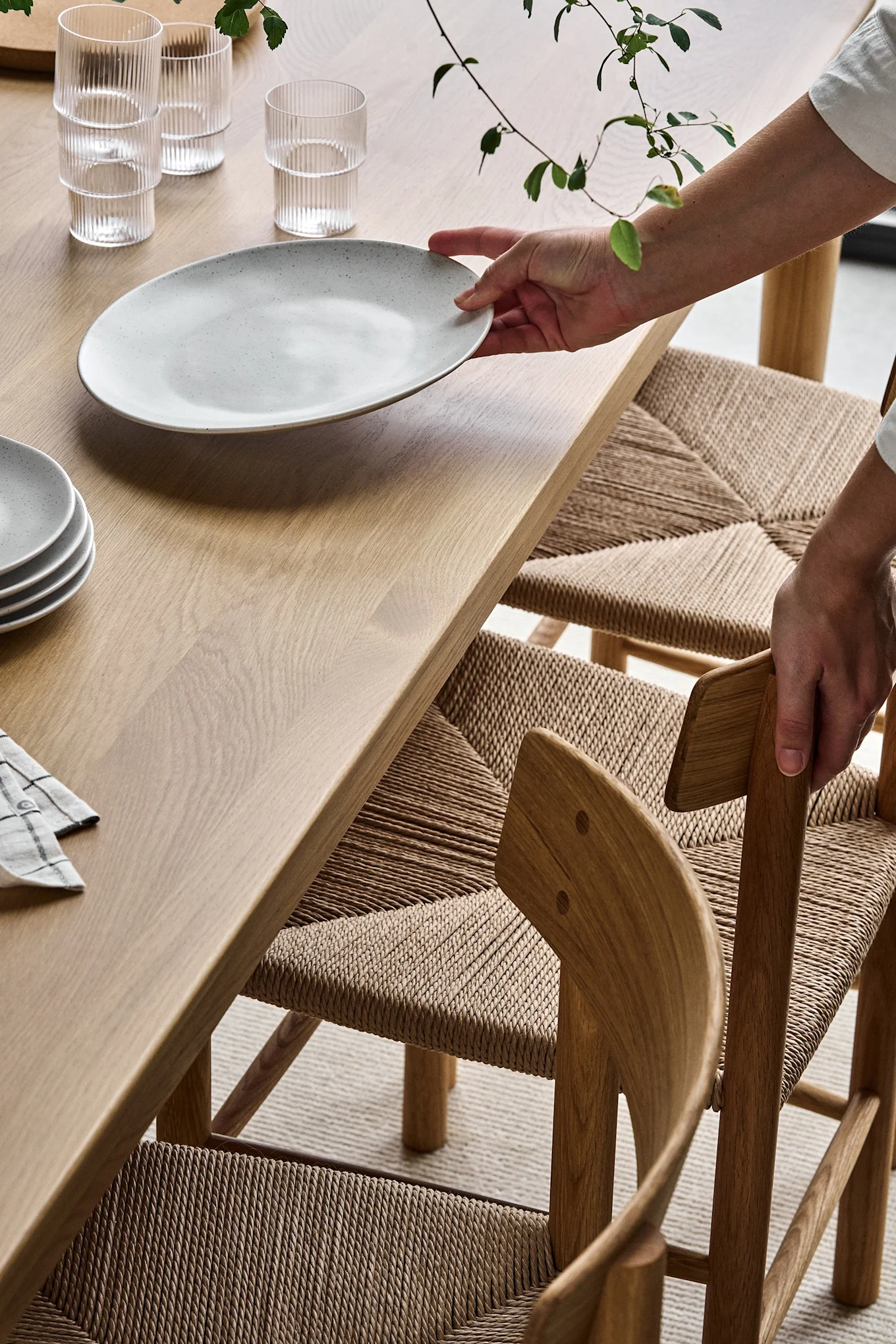 Hands placing a speckled gray plate on a light wooden dining table next to ribbed glasses and woven chairs.