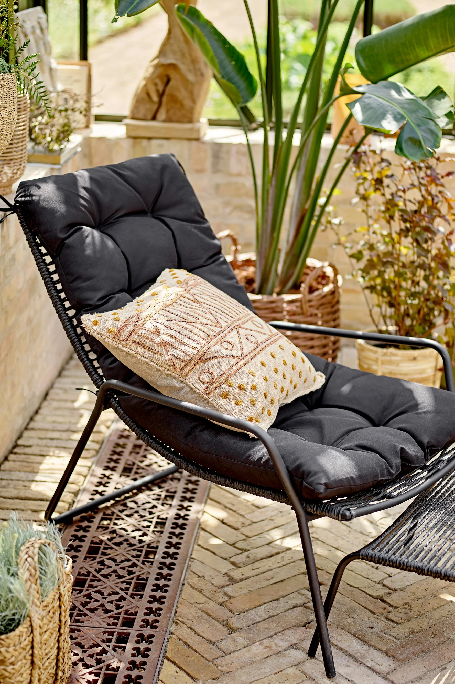 Black lounge chair with dark cushion and patterned beige/gold pillow on a sunny patio with potted plants.
