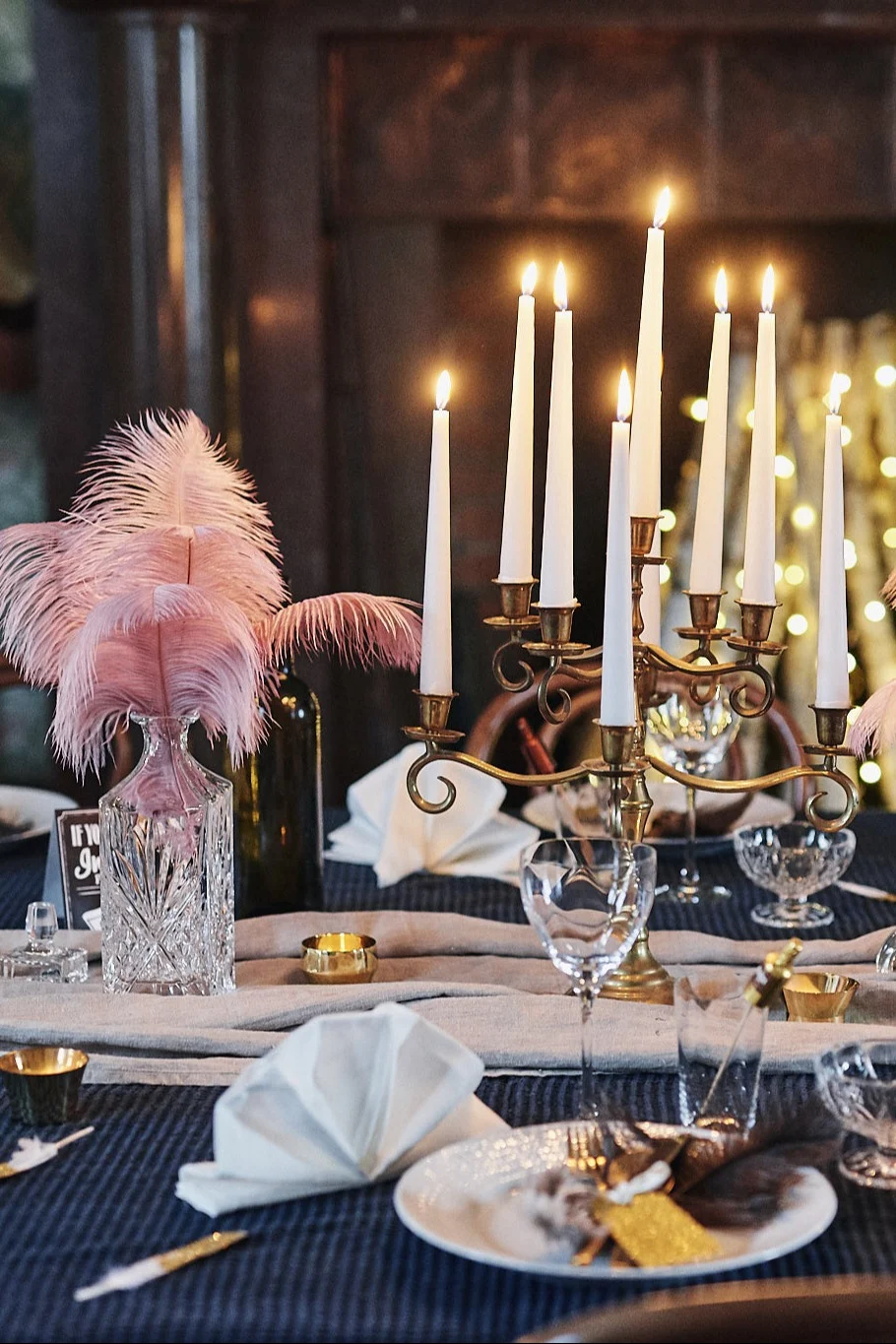 An elegant table setting with a lit brass candelabra, pink feathers in a crystal decanter, and individual place settings with gold accents.