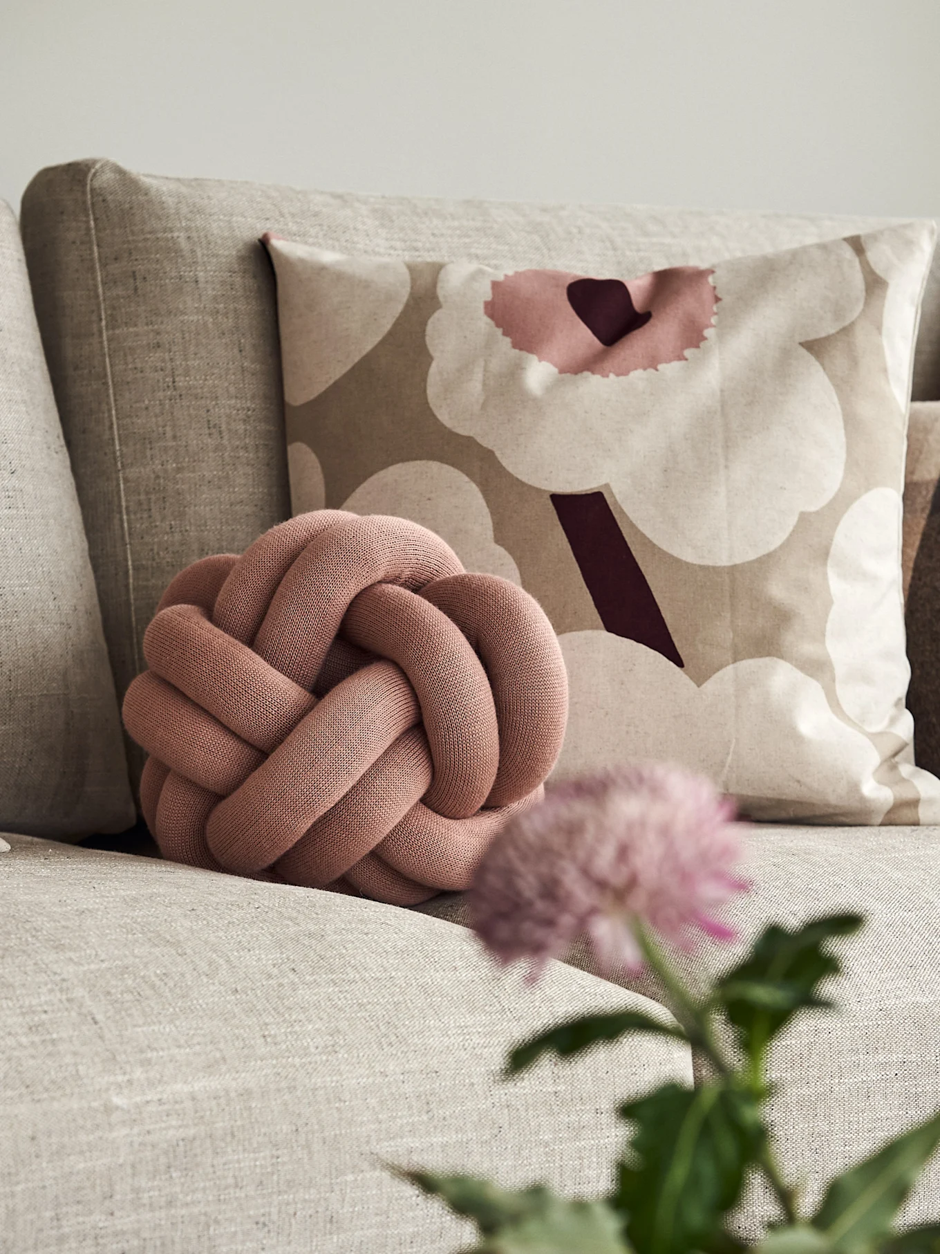 A dusty pink knot cushion sits on a beige couch, next to a patterned cushion with large abstract flowers.