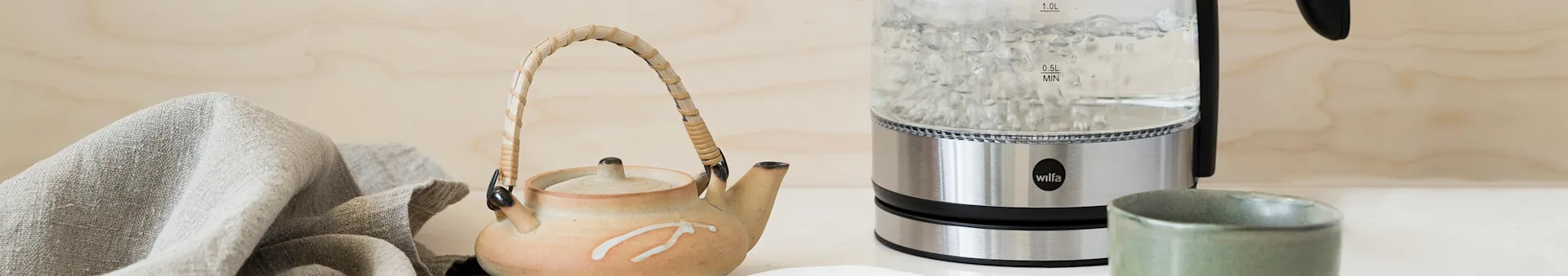 Boiling water in a glass kettle, a ceramic teapot, and a green cup on a countertop with a linen cloth.