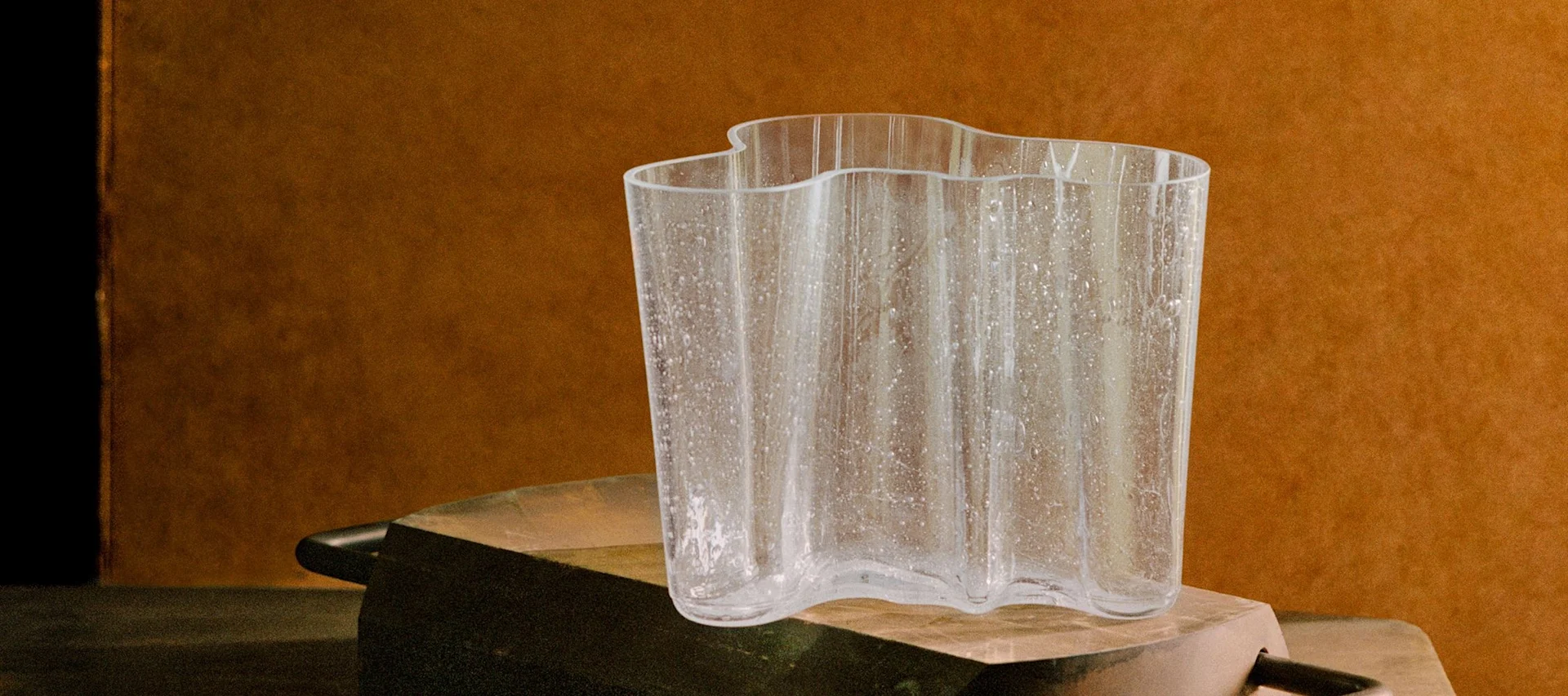 A clear, wavy glass vase with textured speckles on a rustic dark surface against a warm brown background.