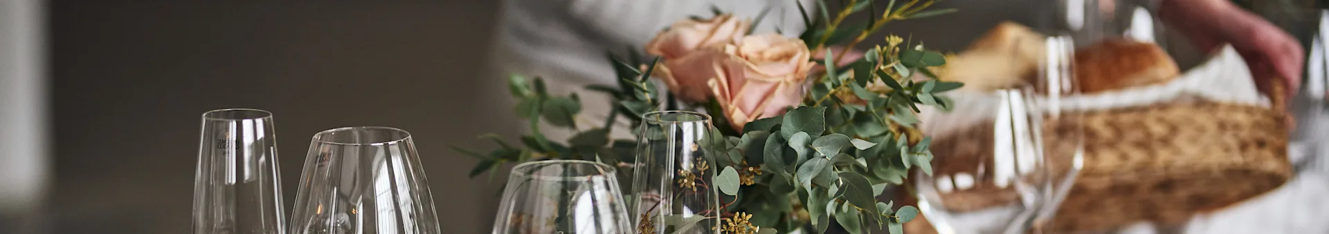 Elegant table setting with clear glasses, pink roses, eucalyptus, and a person holding a bread basket in the background.