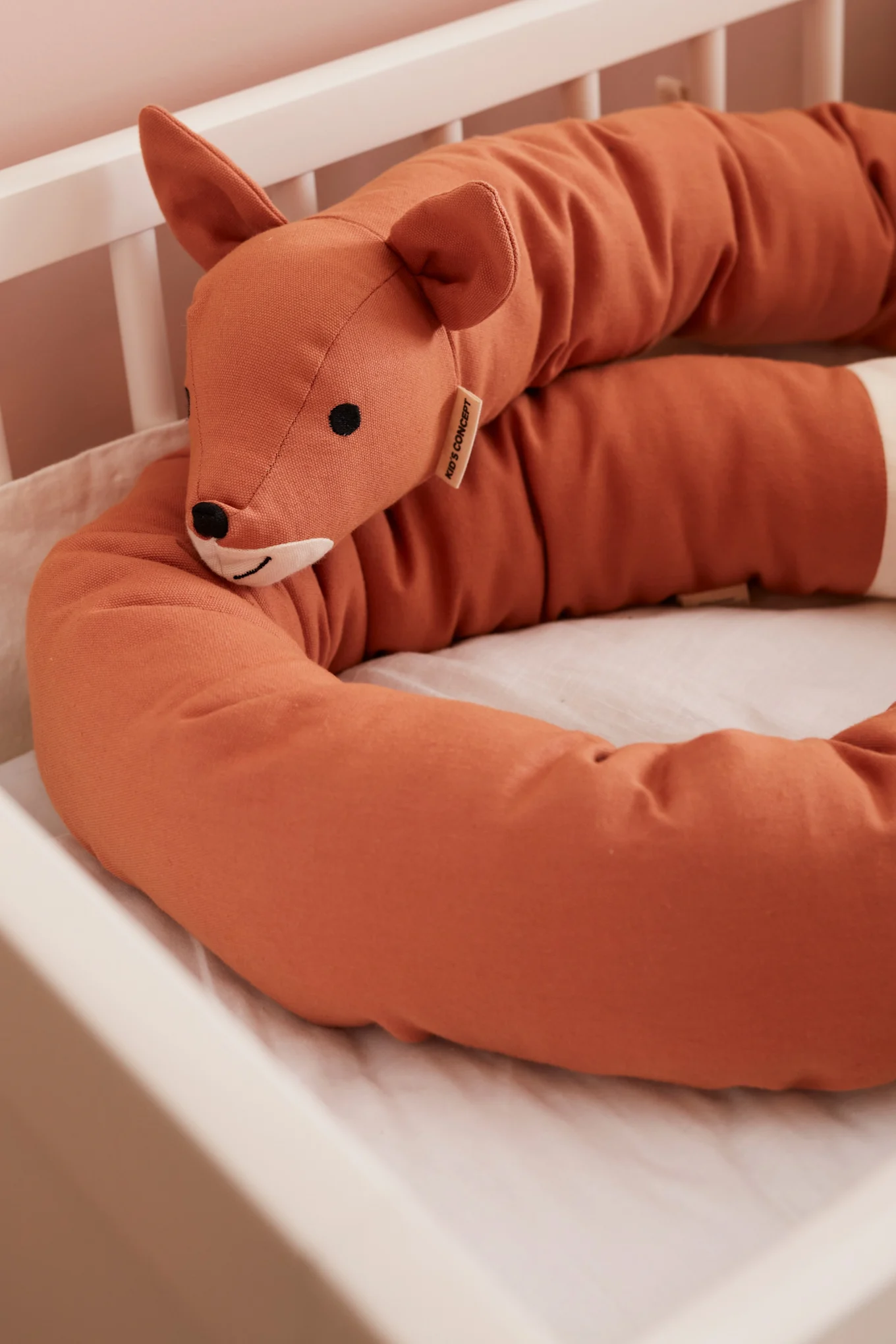 Rust-orange fox-shaped crib bumper with a 'KID'S CONCEPT' tag, resting inside a white baby crib.