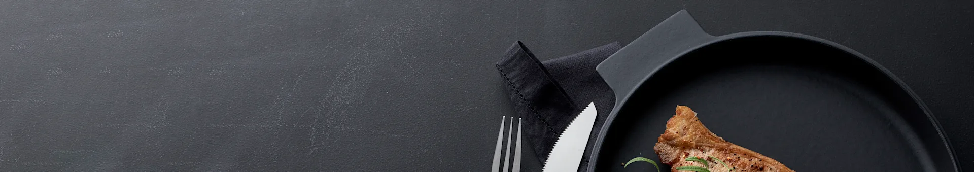 Roasted meat chop with rosemary and pepper on a black plate, with a fork and serrated knife on a dark background.